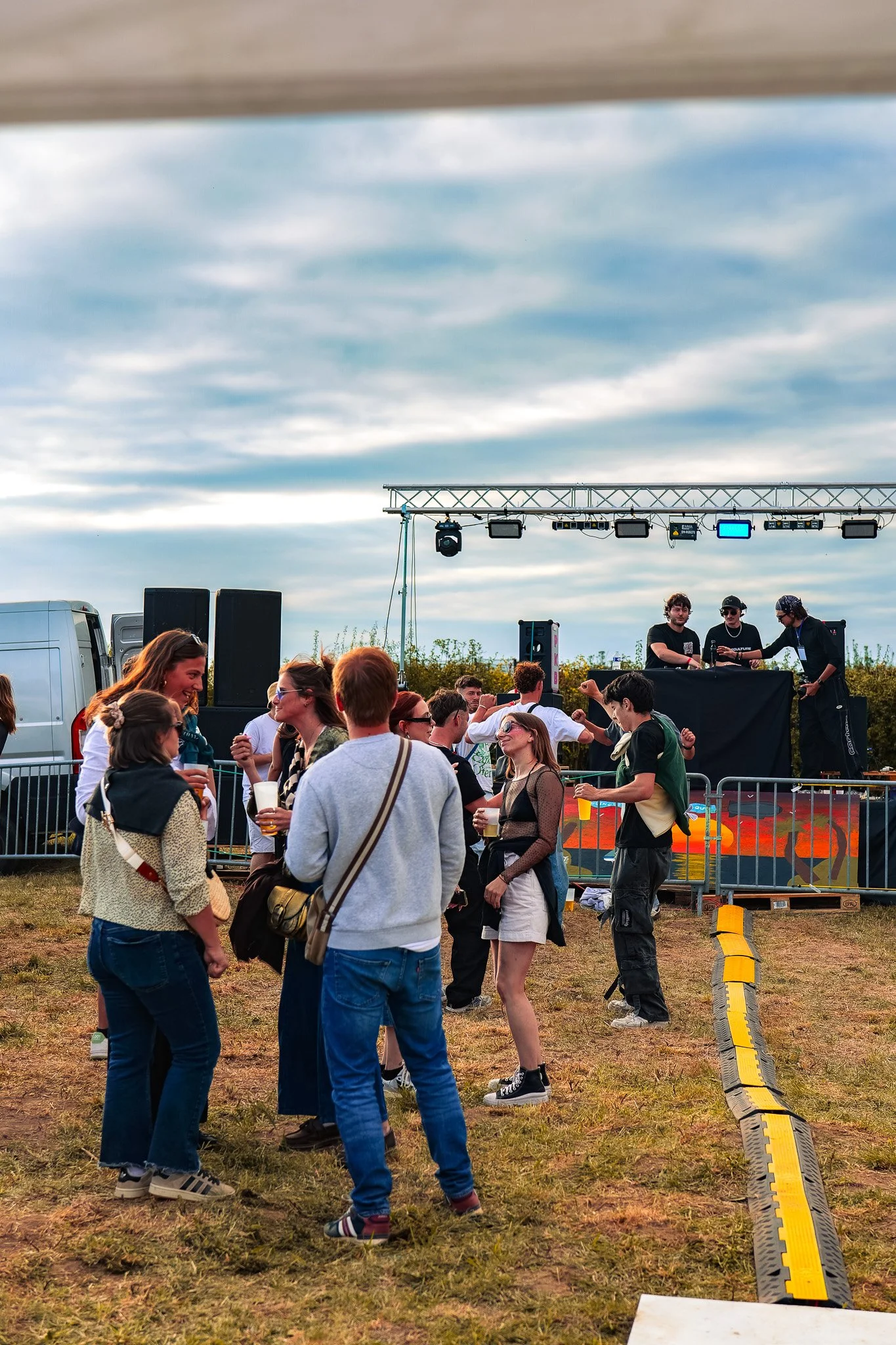 Groupe de personnes debout devant une scène de festival avec un toit, musique jouée par un DJ derrière. Les gens sourient, discutent et tiennent des boissons. Le ciel est nuageux, l'ambiance est détendue et festive.