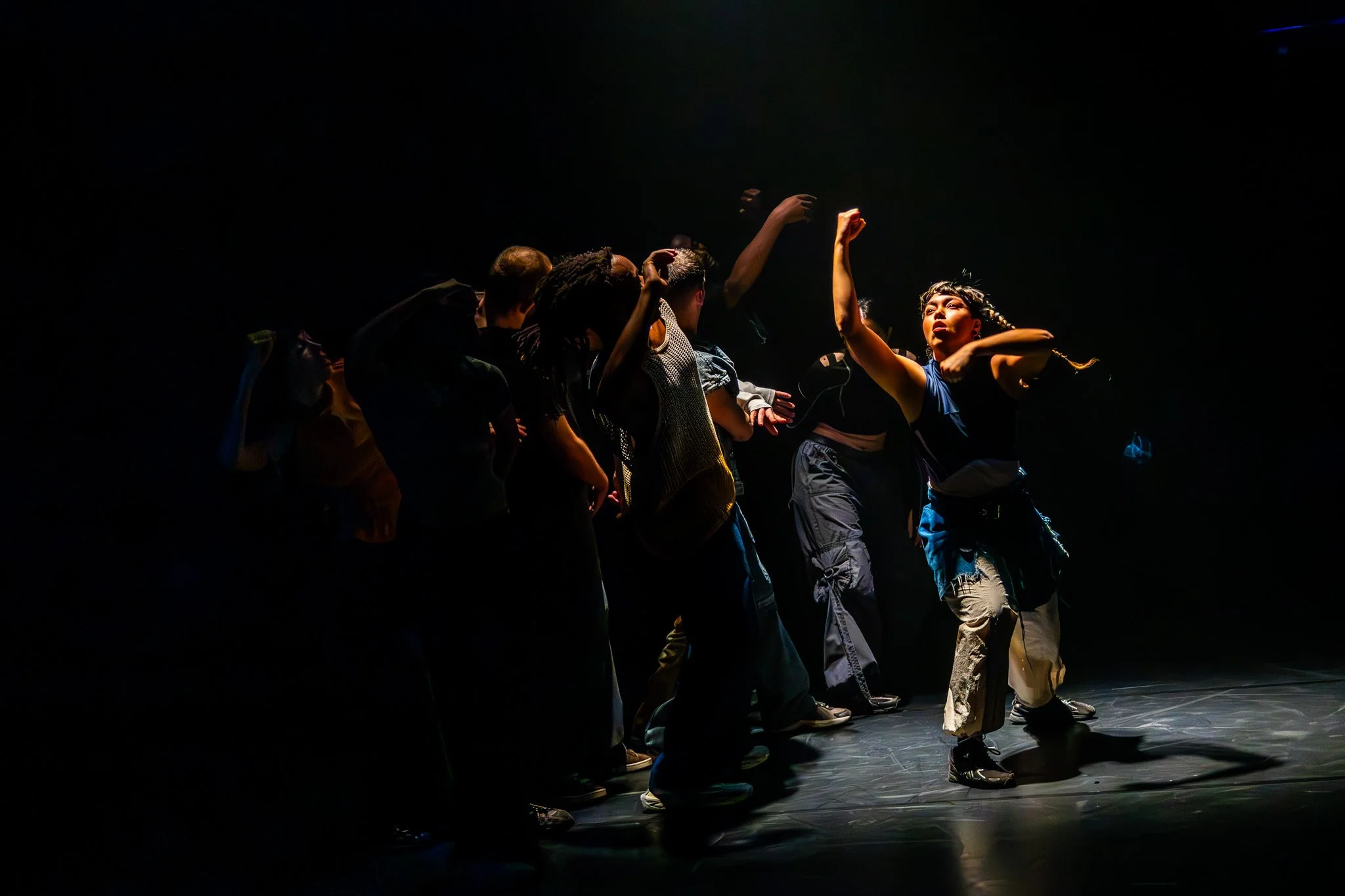 Groupe de danseurs en pleine performance sur scène, éclairés par une lumière focalisée dans l'obscurité.