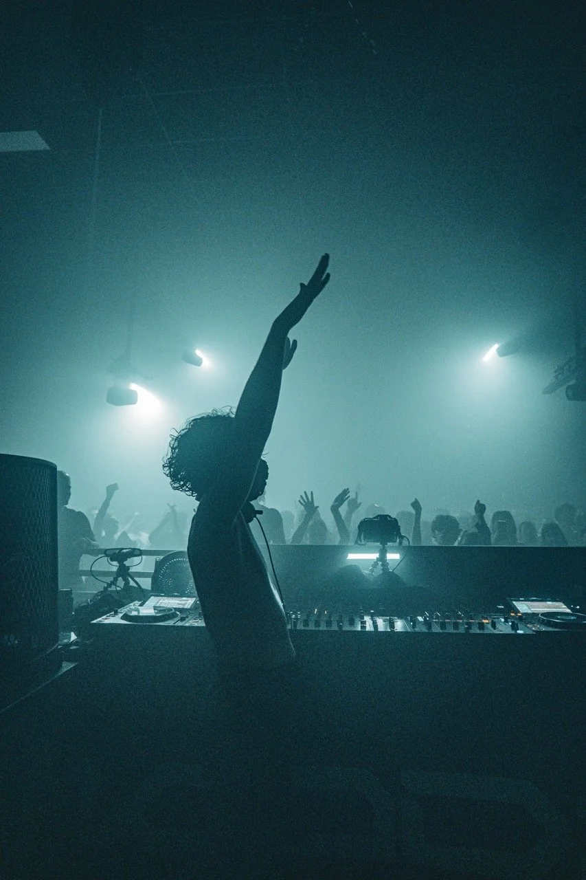 DJ avec une main levée, en train de mixer dans une boîte de nuit sombre avec des lumières et un public qui danse en arrière-plan.