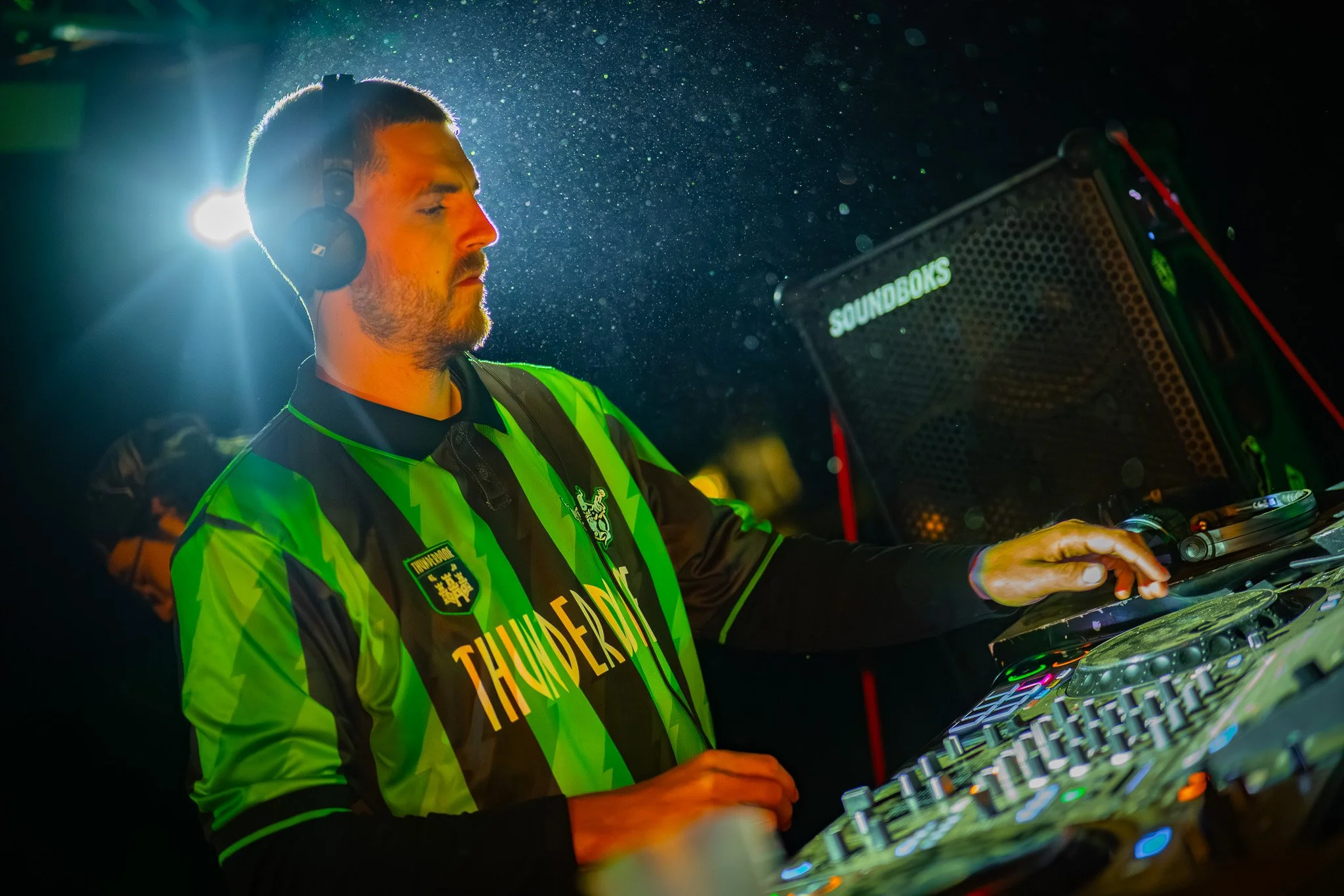 Un DJ porte un casque, joue avec des équipements de musique, devant un arrière-plan avec des particules de poussière ou de pluie.