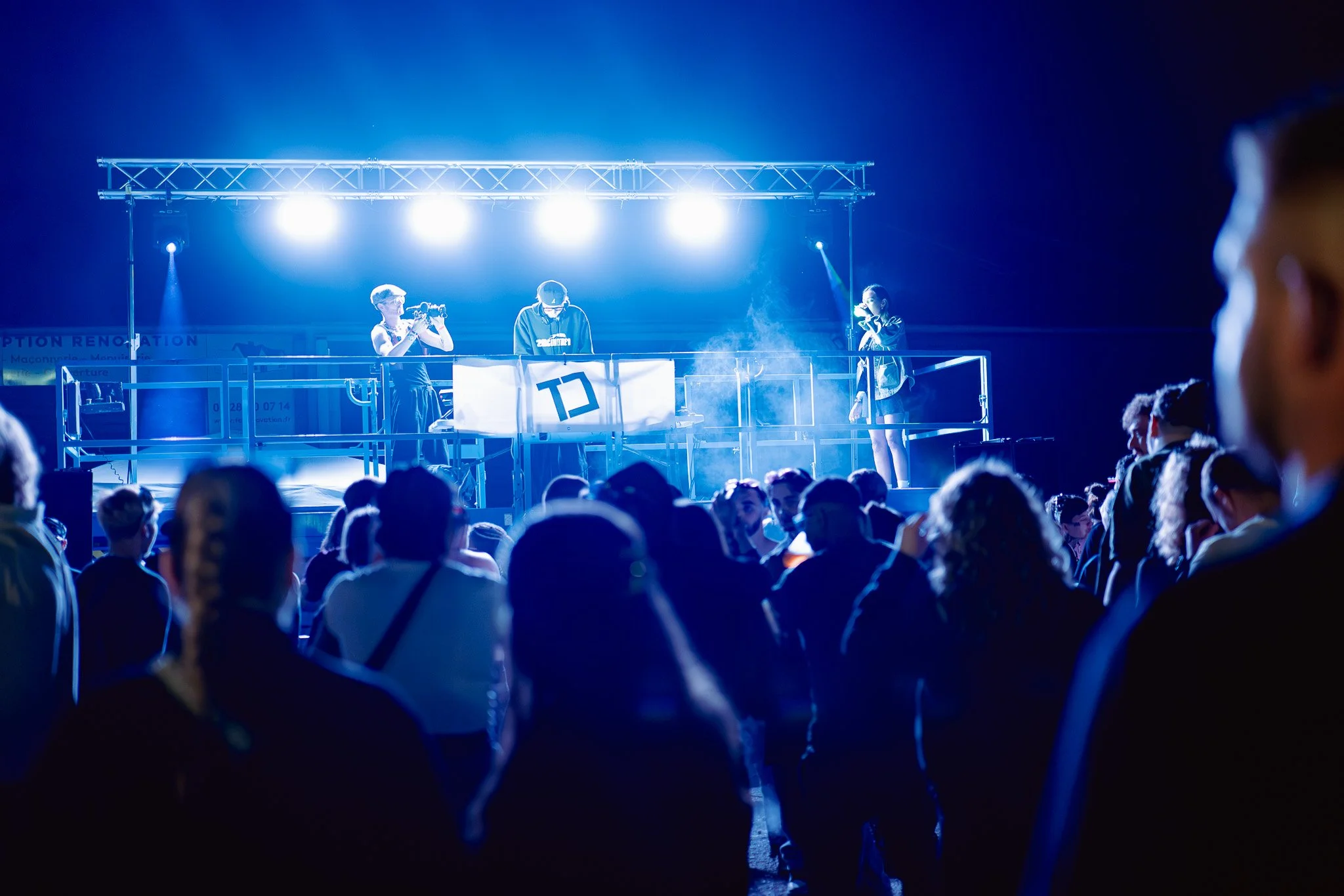 Un groupe de personnes assises dans une salle ou un auditorium, regardant un groupe de DJ en train de jouer sur une scène éclairée par de fortes lumières, dans une ambiance de concert ou de festival nocturne.