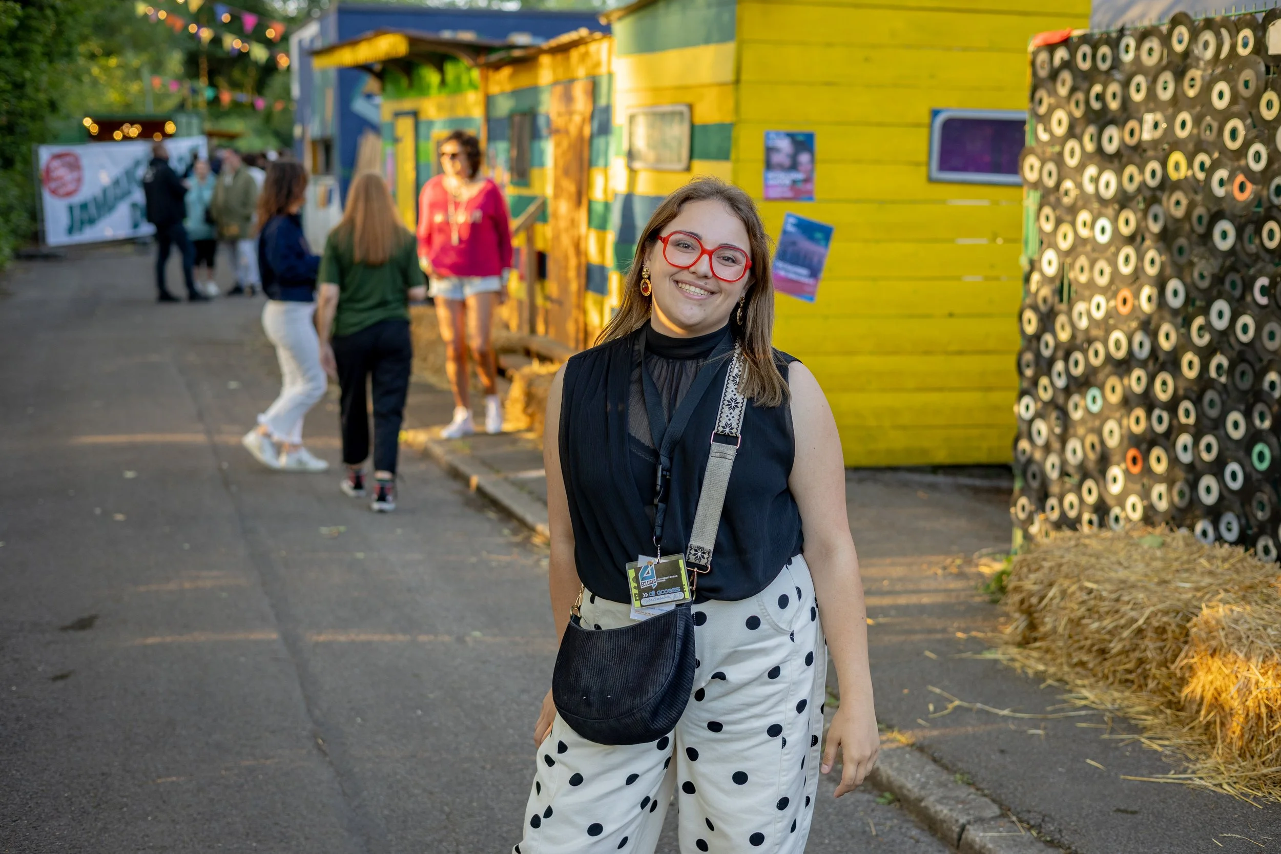 Une femme souriante portant des lunettes rouges, une jupe à pois, et un badge, se tient devant une structure colorée lors d'un événement extérieur en soirée.