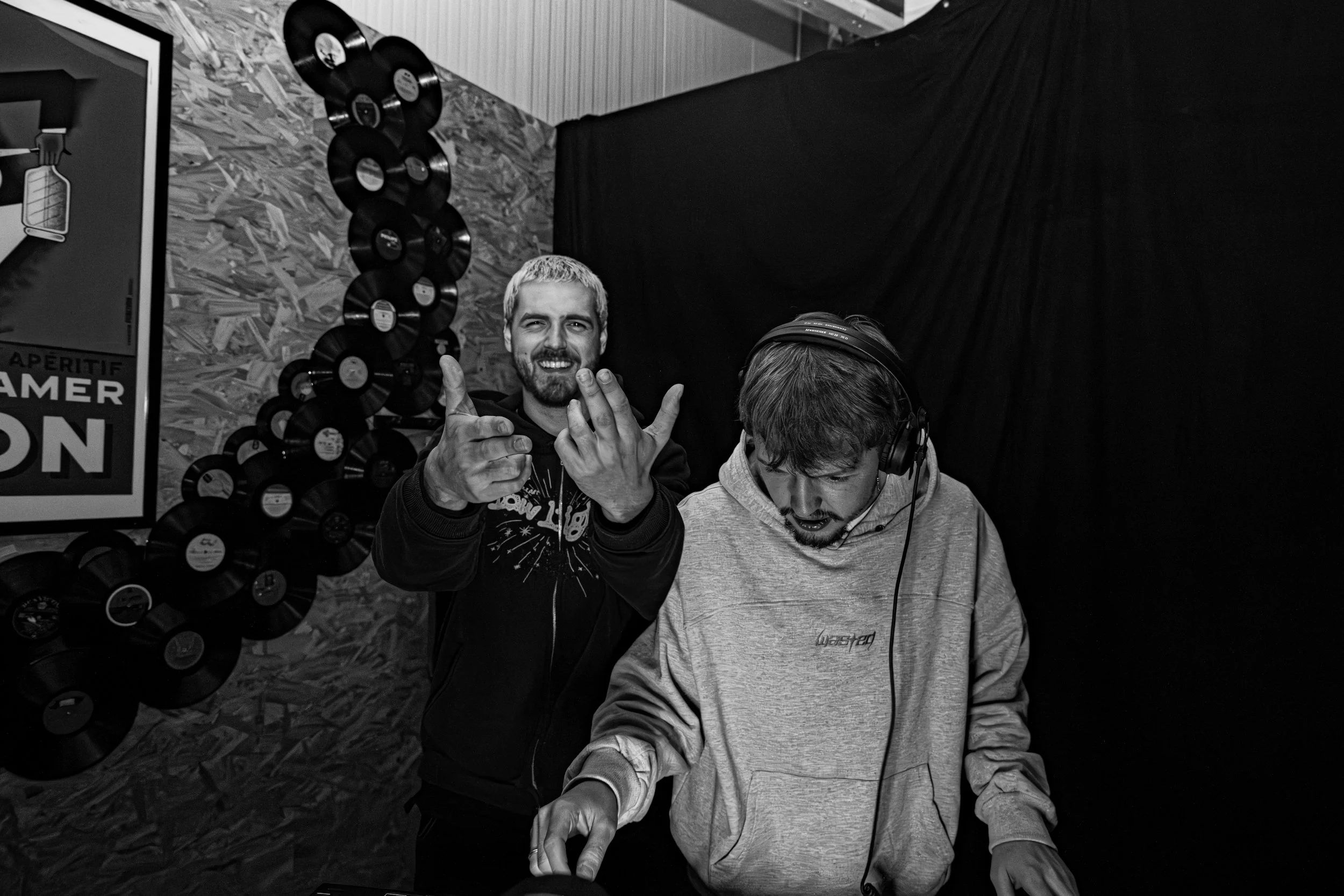Deux jeunes hommes dans une ambiance détendue, avec un décor de disques vinyles au mur, l'un souriant et faisant un geste de paix avec les doigts, l'autre concentré avec des écouteurs, dans un cadre intérieur