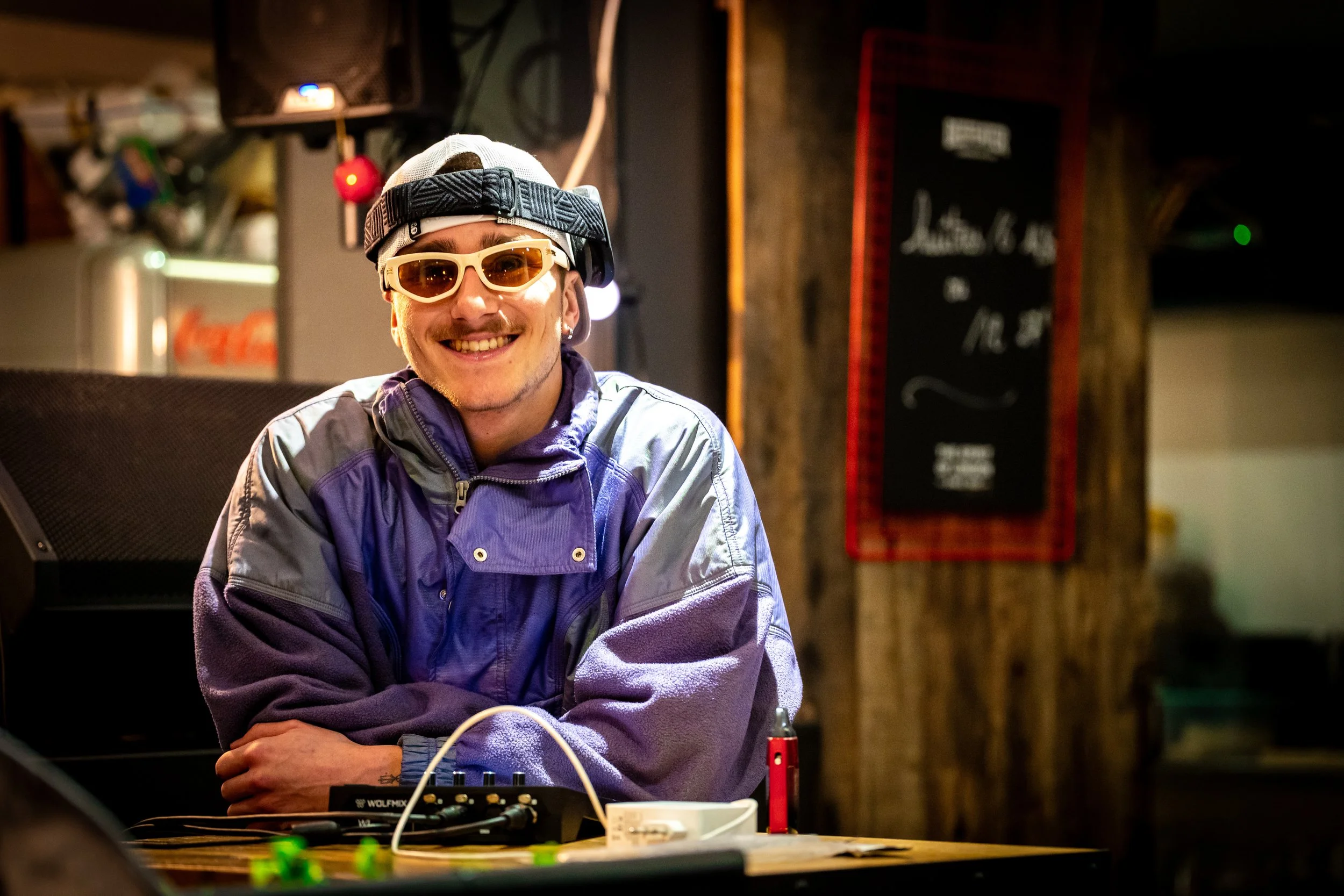 Un homme souriant porte des lunettes de soleil jaunes, un chapeau gris, et un veston violet, assis à une table avec du matériel électronique, dans un lieu intérieur avec un panneau noir en arrière-plan.