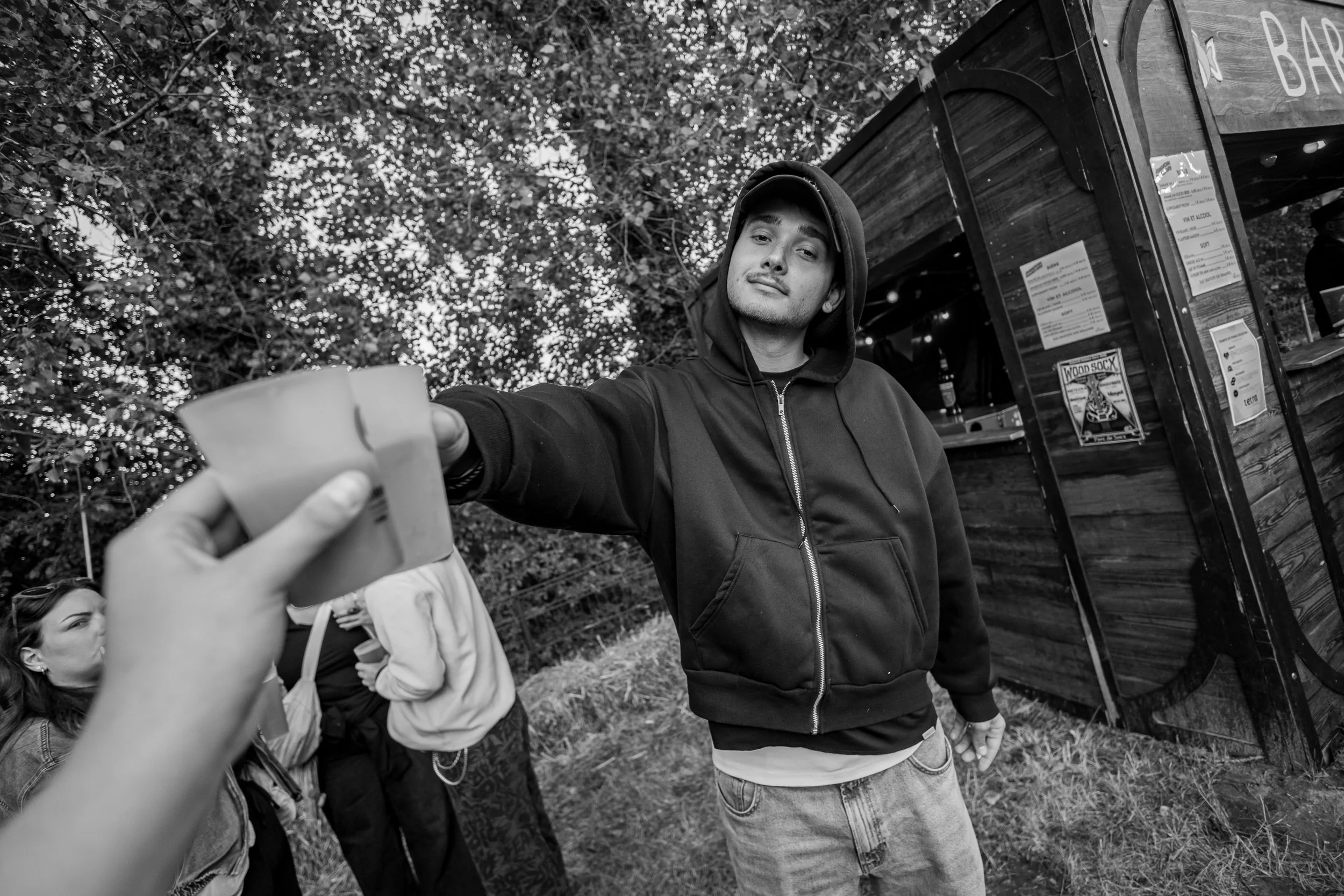 Un jeune homme portant une veste à capuche et un pantalon en denim, tenant un verre à la main, se trouve de face devant un stand en bois avec un panneau de menu, lors d'une occasion en plein air, avec des arbres en arrière-plan et plusieurs autres pe