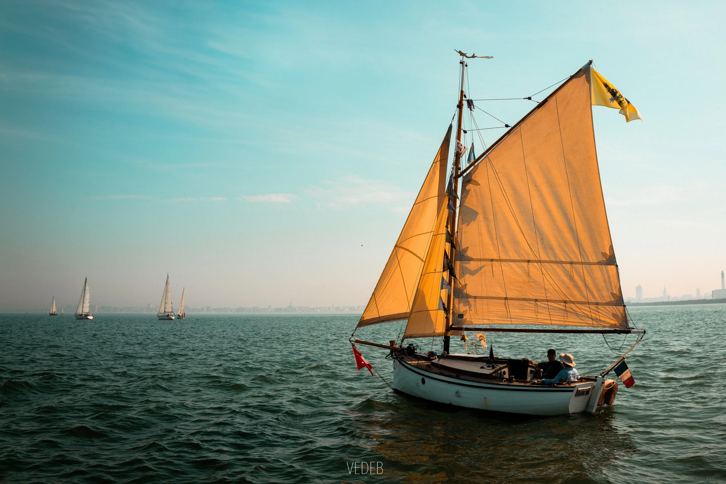 Un voilier avec voiles orange voguant sur une mer calme, avec d'autres voiliers en arrière-plan et une ligne d'horizon avec une tour au loin. Ciel clair avec quelques nuages.