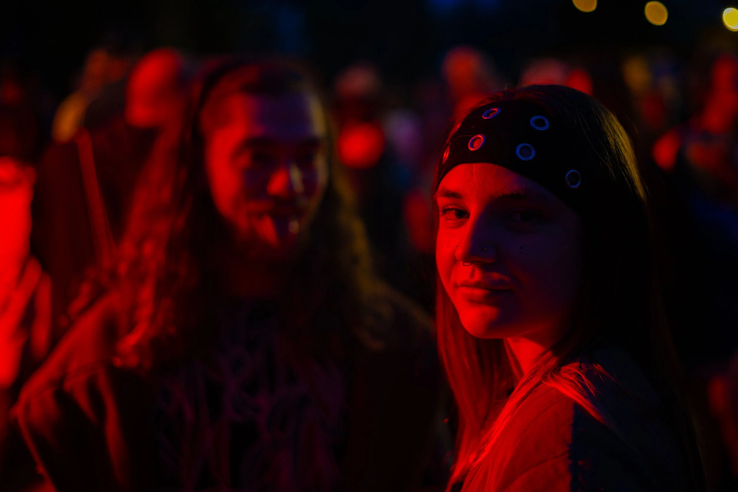 Groupe de personnes dans une fête ou un concert, éclairage tamisé avec des lumières colorées, un jeune homme et une jeune femme en premier plan, la femme porte un bandeau lumineux.