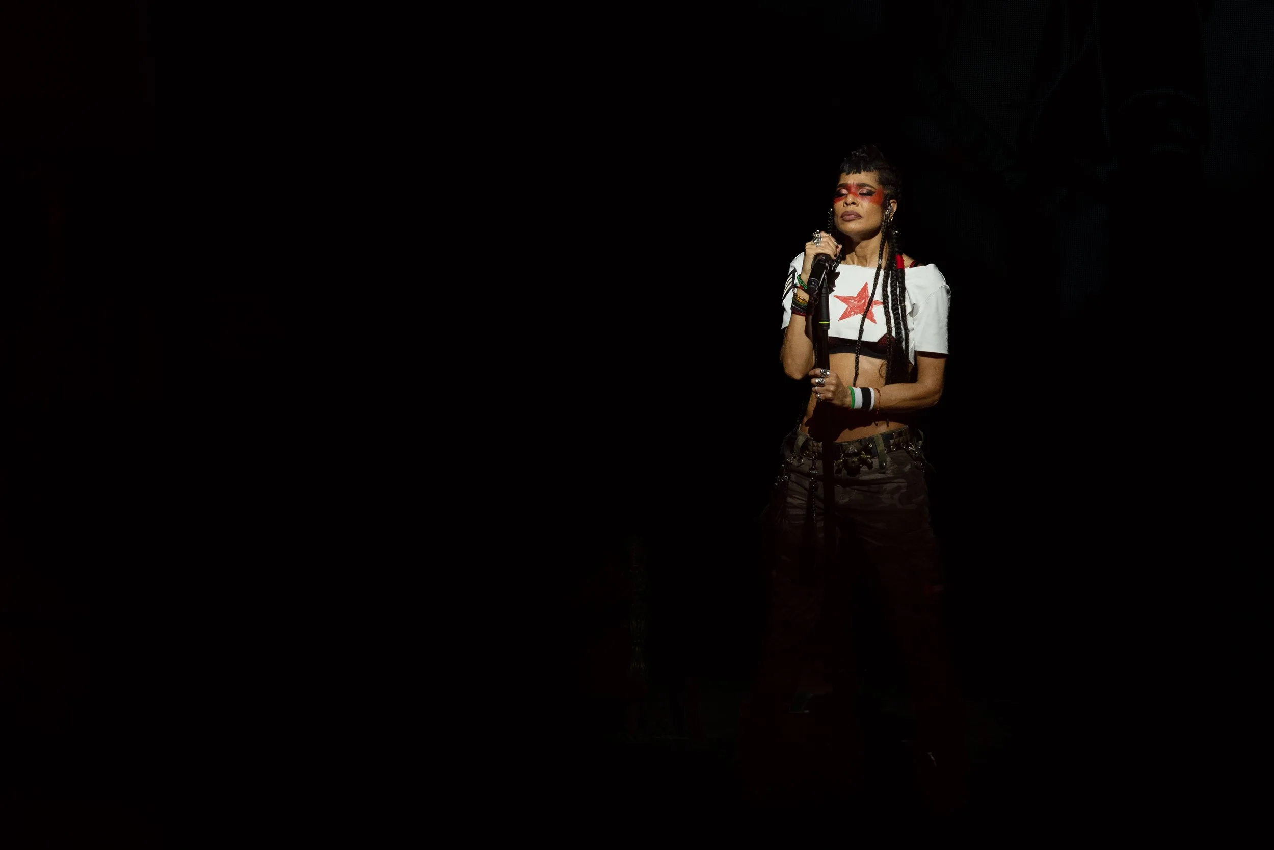 Une femme chante sur scène, habillée d'un crop top blanc avec une étoile rouge, porte des accessoires colorés, avec un fond noir.