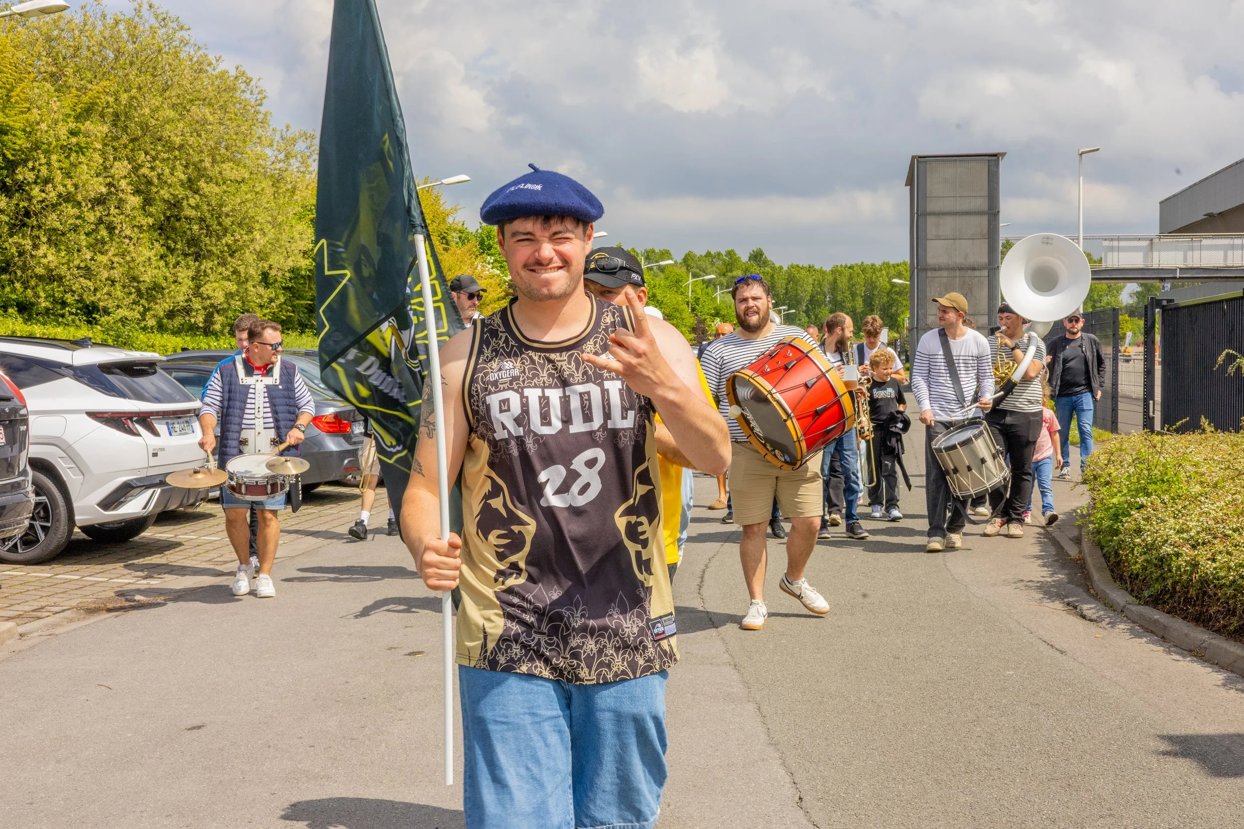 Un groupe de personnes marchant dans une rue, parmi elles un homme souriant portant un béret bleu, des instruments de musique et un drapeau, lors d'une parade ou d'un festival en plein air