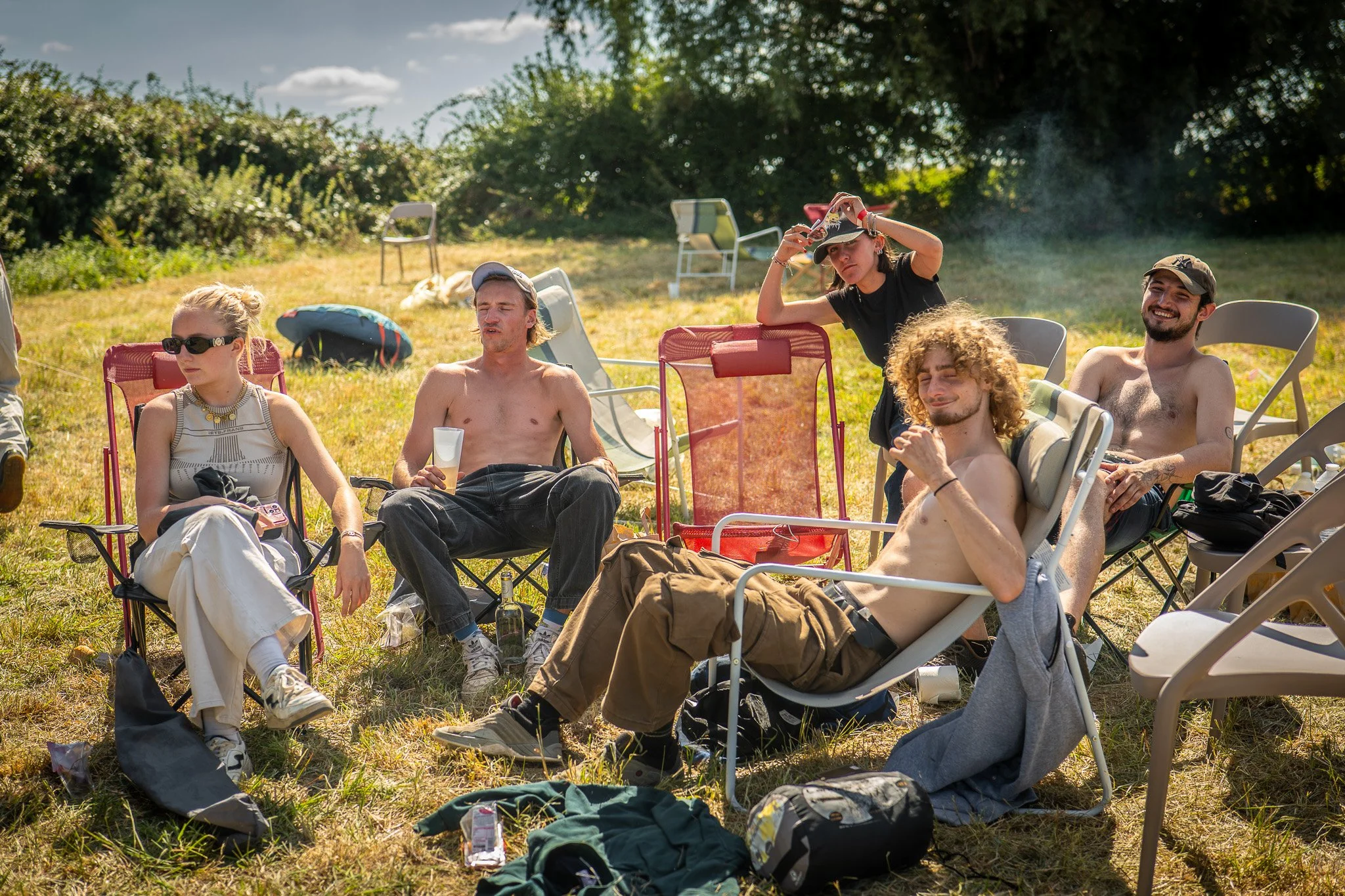Groupe de jeunes adultes assis et allongés dans un champ, certains en maillot de bain, profitant d'un moment ensoleillé en plein air avec des chaises pliantes et des sacs autour.