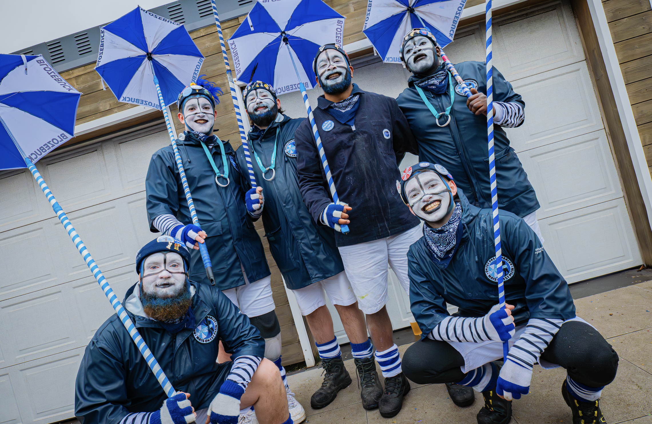 Groupe de personnes habillées en costumes de mime avec maquillage blanc, portant des parapluies bleus et blancs avec l'inscription 'BROSEBRUCK', posant devant un garage en bois