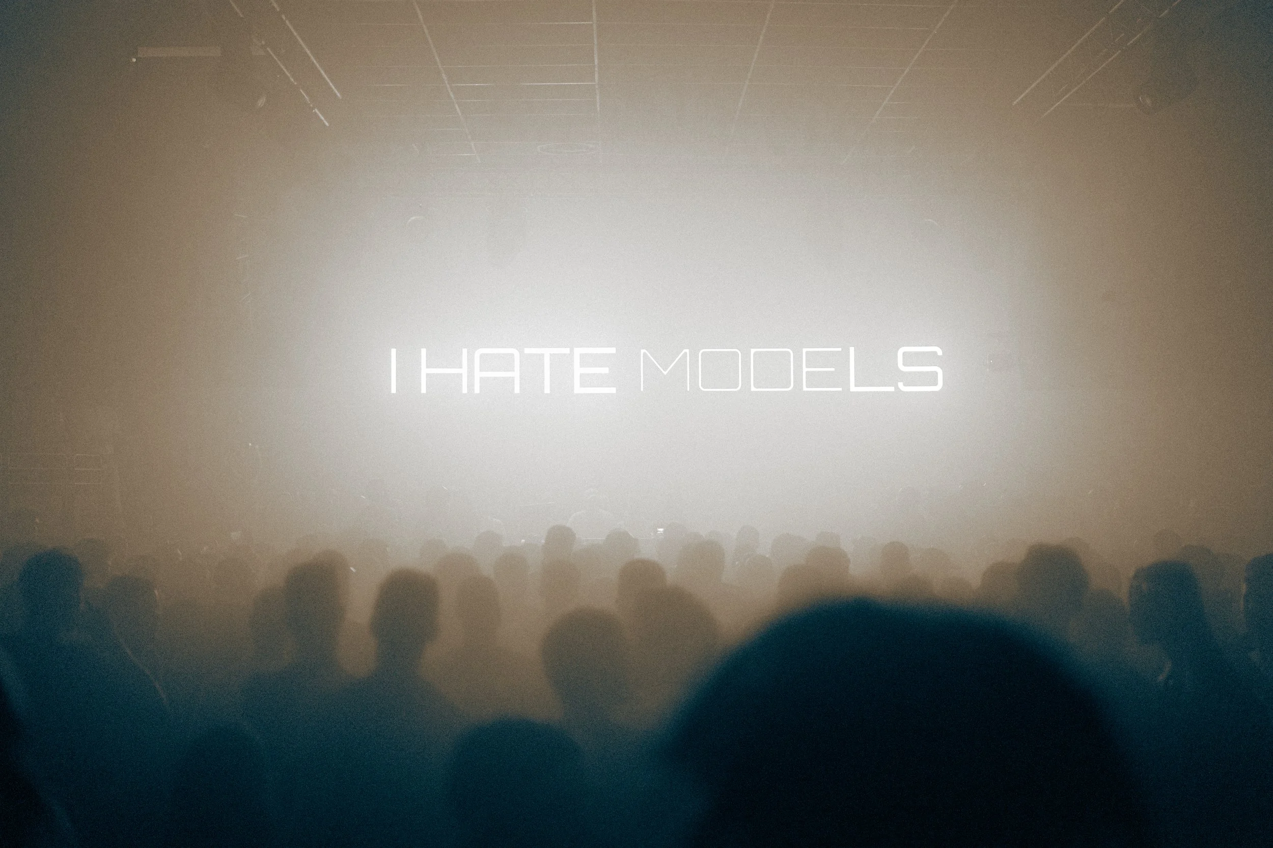 Une scène sombre avec une lumière brillante au centre affichant le texte 'I HATE MODELS' avec une audience assise face à l'écran.