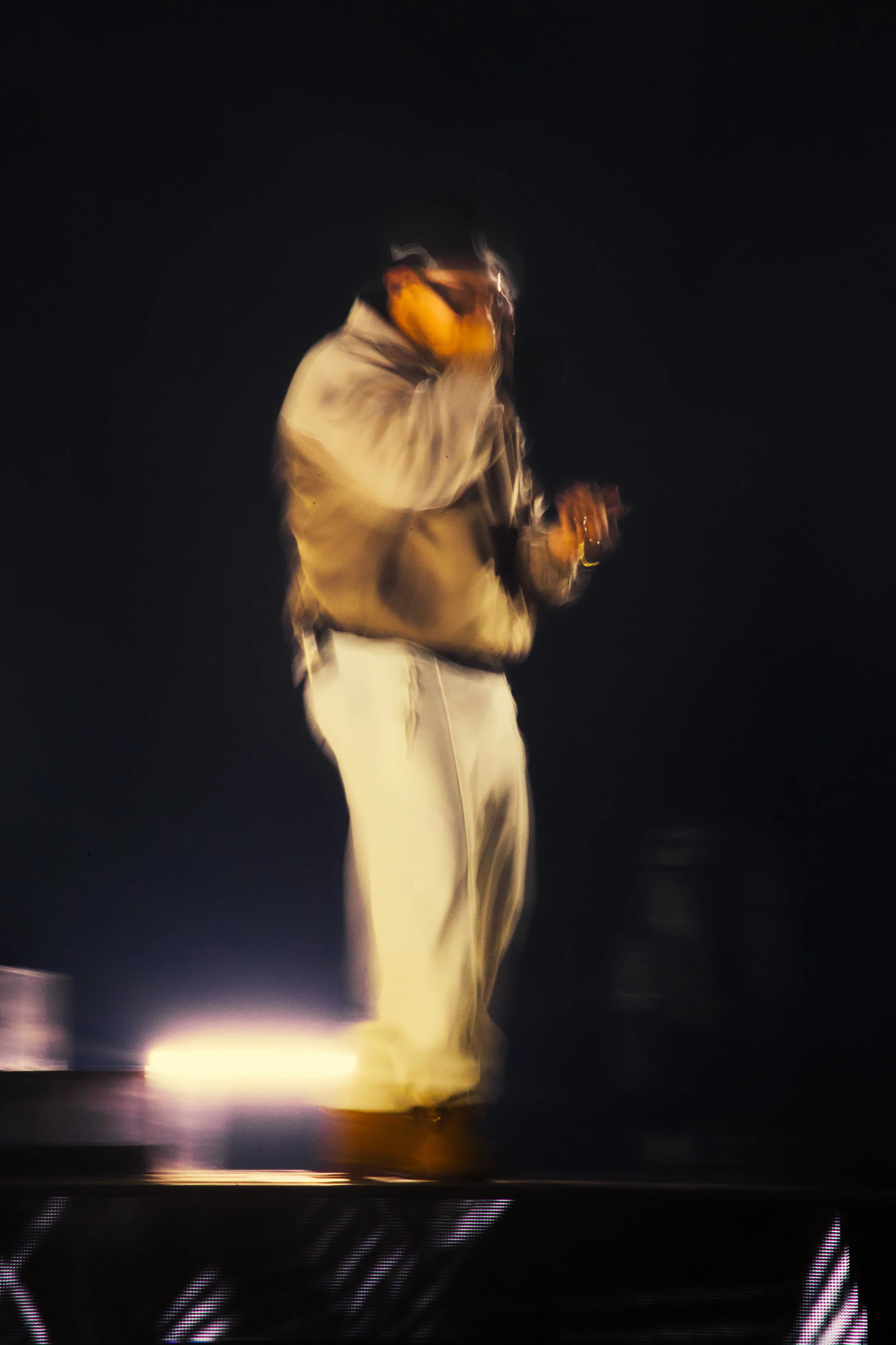Person portant une veste beige et des pantalons beige clair, tenant un objet dans la main, dans un environnement sombre.