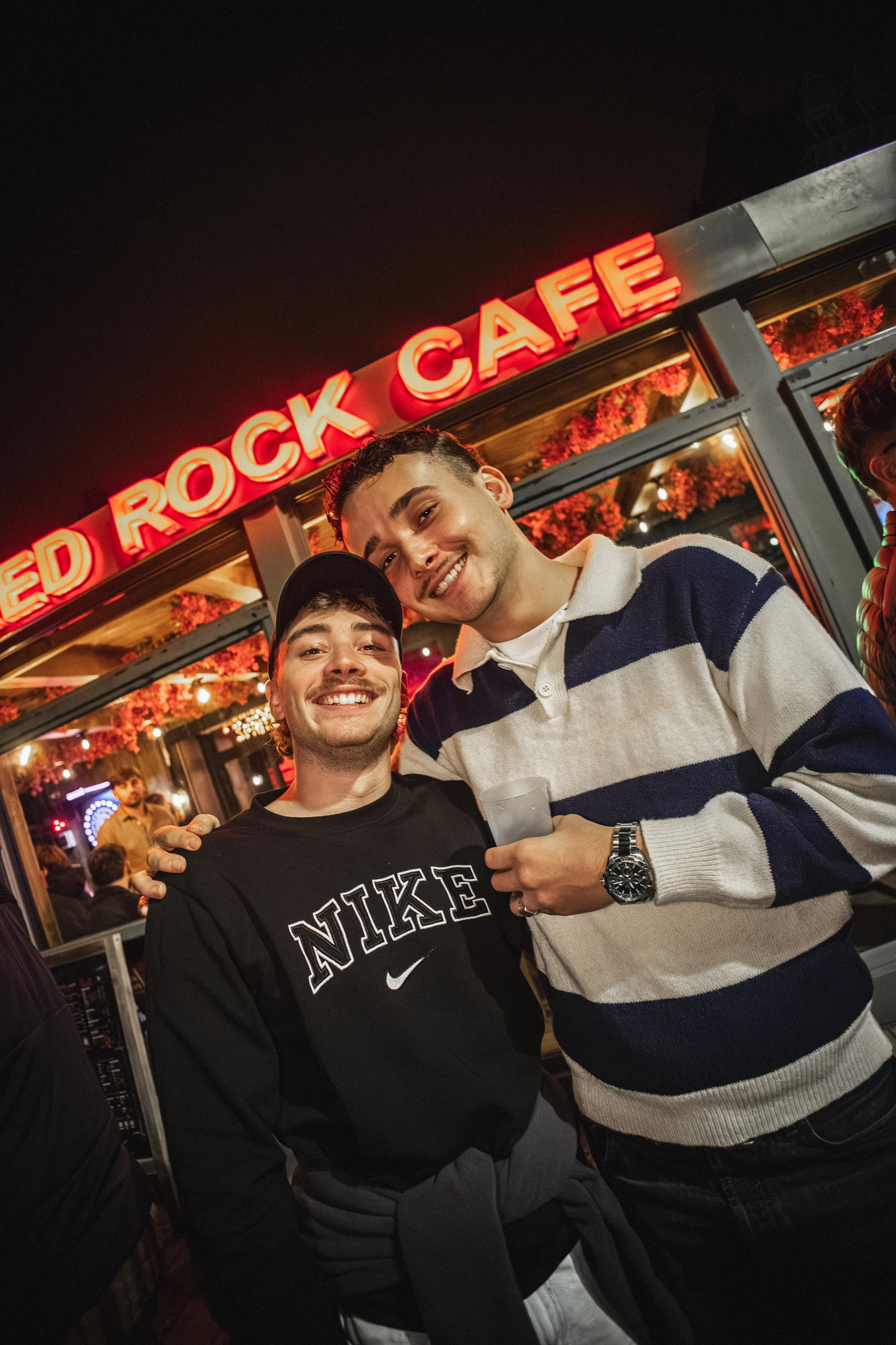 Deux jeunes hommes souriants posent ensemble devant une enseigne lumineuse rouge à l'extérieur d'un restaurant ou un bar nocturne.
