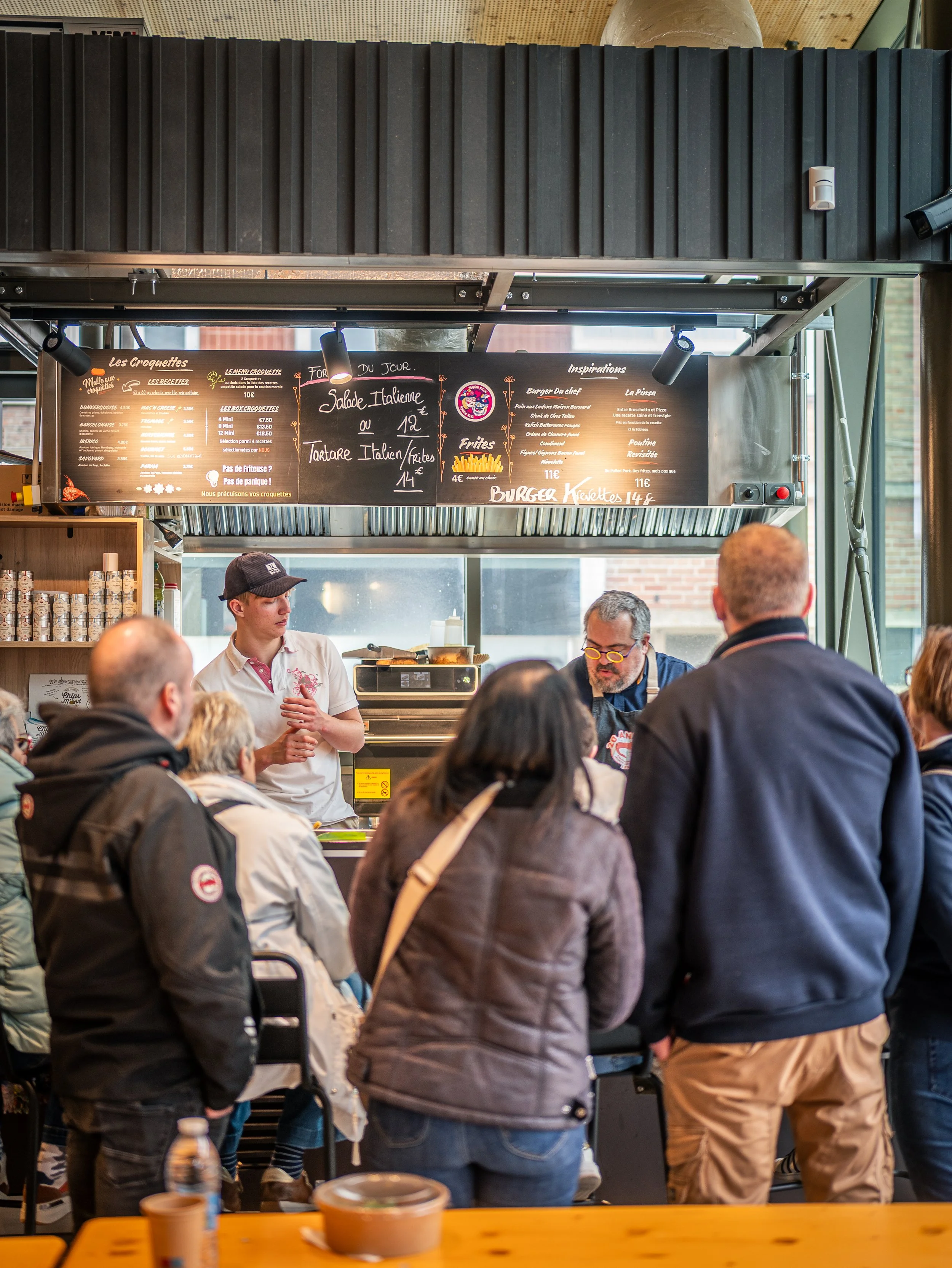Une scène dans un restaurant ou un café avec plusieurs personnes attendent ou commandent de la nourriture, un menu écrit à la craie est visible au fond avec des plats italiens.