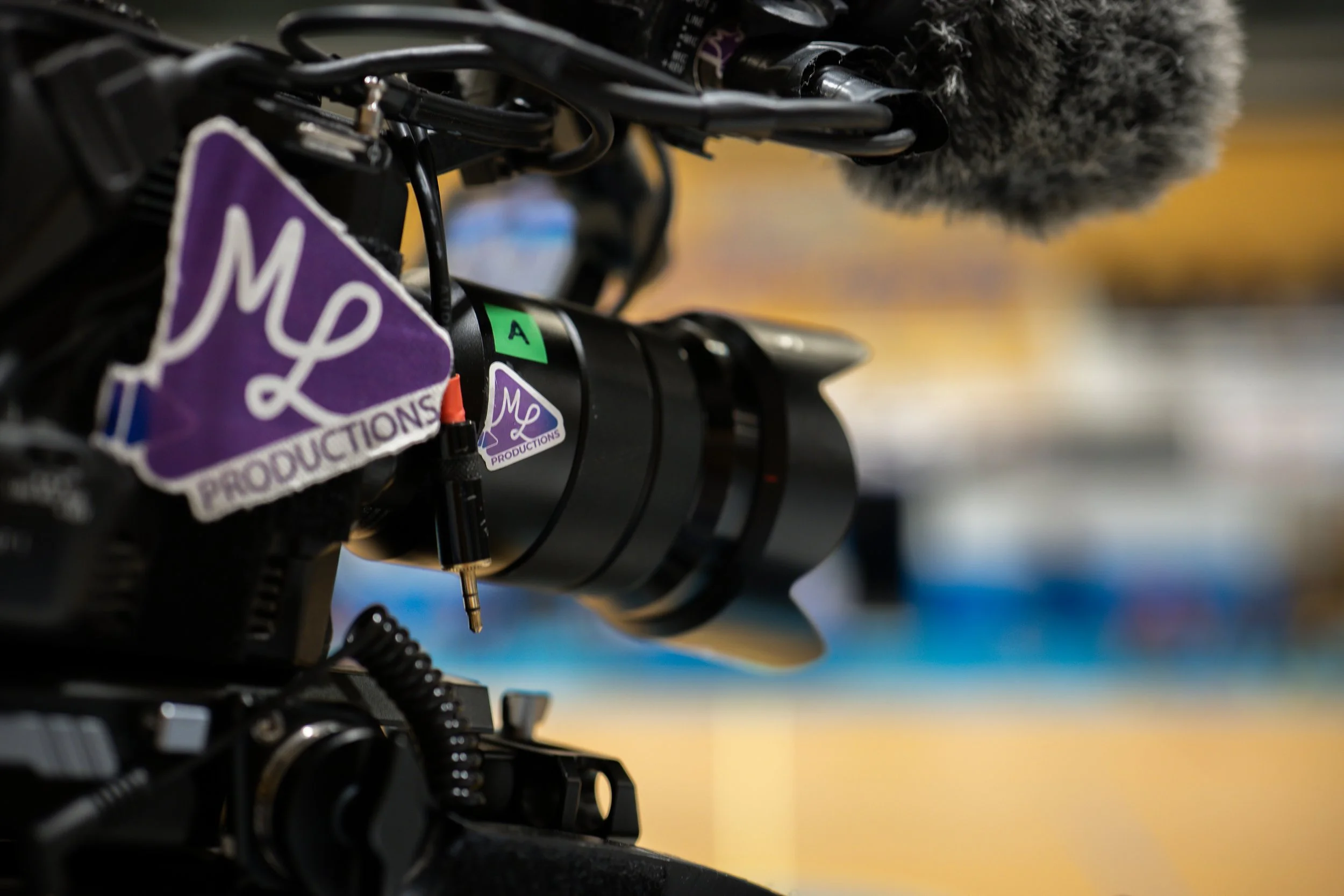 Plan rapproché d'une caméra ou d'un équipement de production vidéo avec des câbles noirs, un microphone à soufflet gris et des autocollants violets avec des lettres, en arrière-plan flou jaune et bleue.