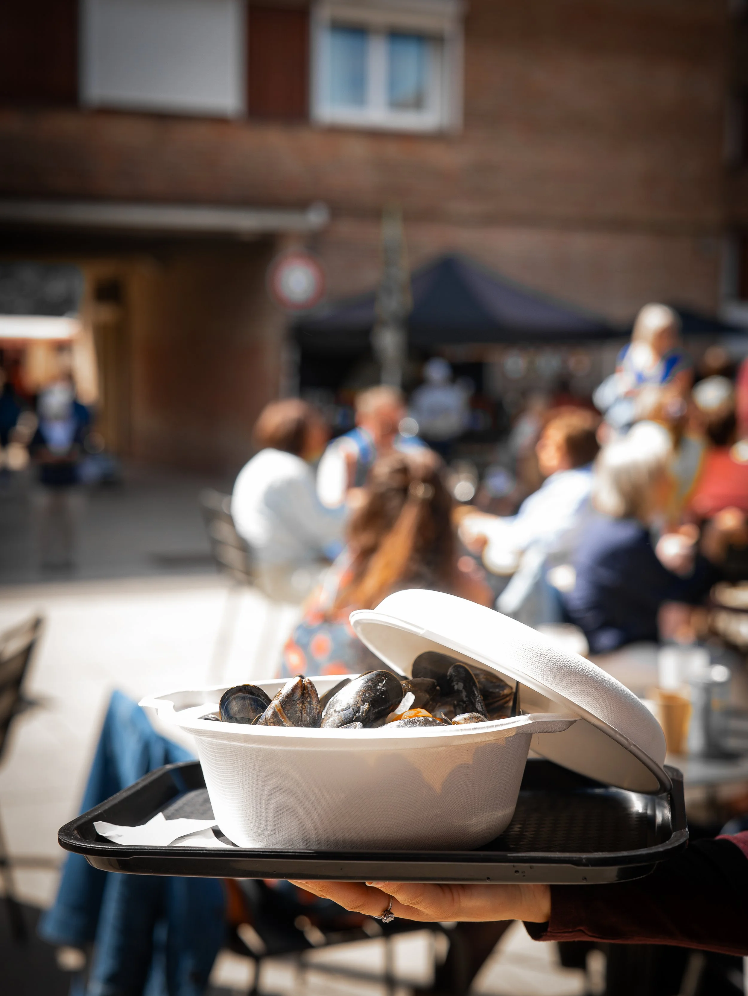 Un plateau avec des moules sur une table lors d'un événement en plein air; fond flou avec des personnes et une bâtisse en brique.