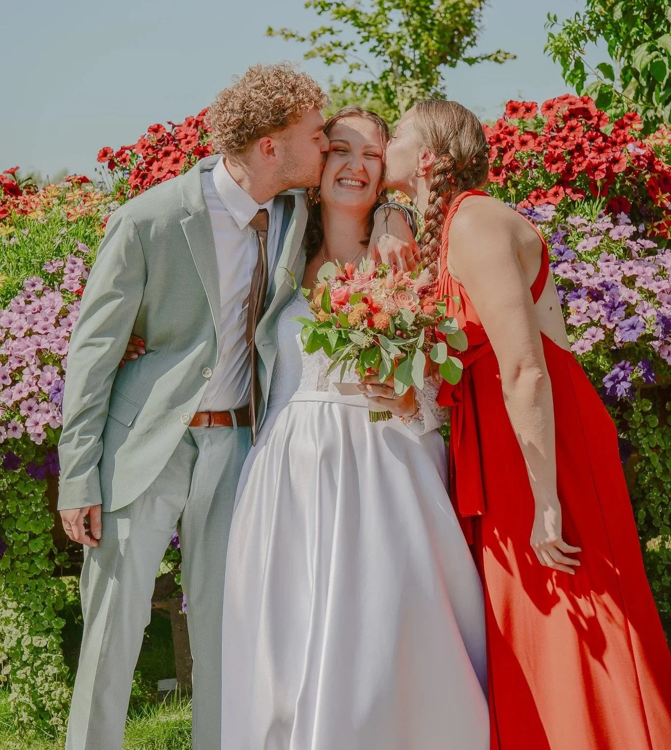 Un couple de mariés avec une femme tenant un bouquet de fleurs, un homme et une femme l'embrassant sur les joues, entourés de fleurs colorées dans un jardin.