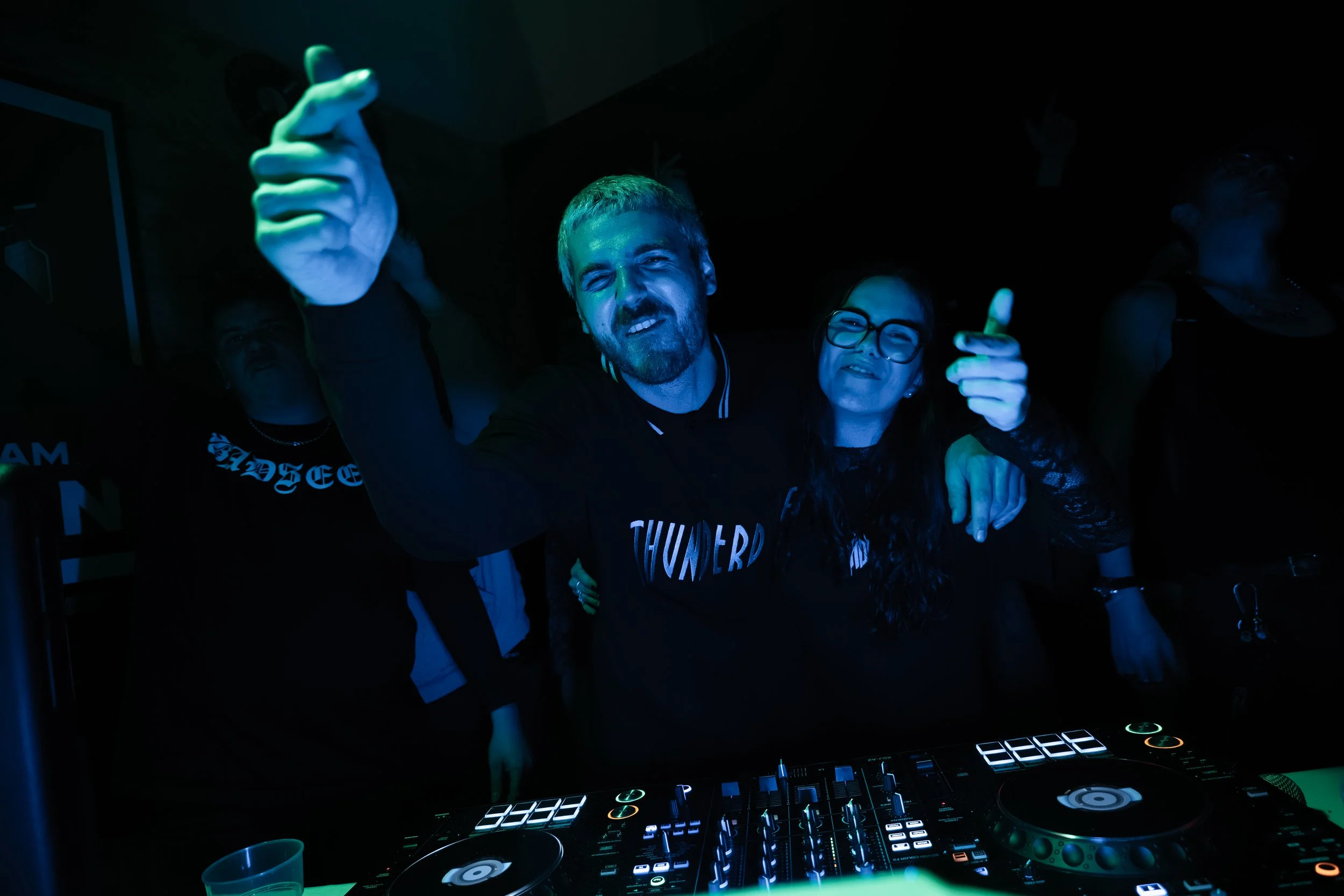Groupe de personnes dans une soirée, un homme est en train de mixer de la musique avec un DJ contrôleur, ambiance sombre avec des lumières vertes et bleues.