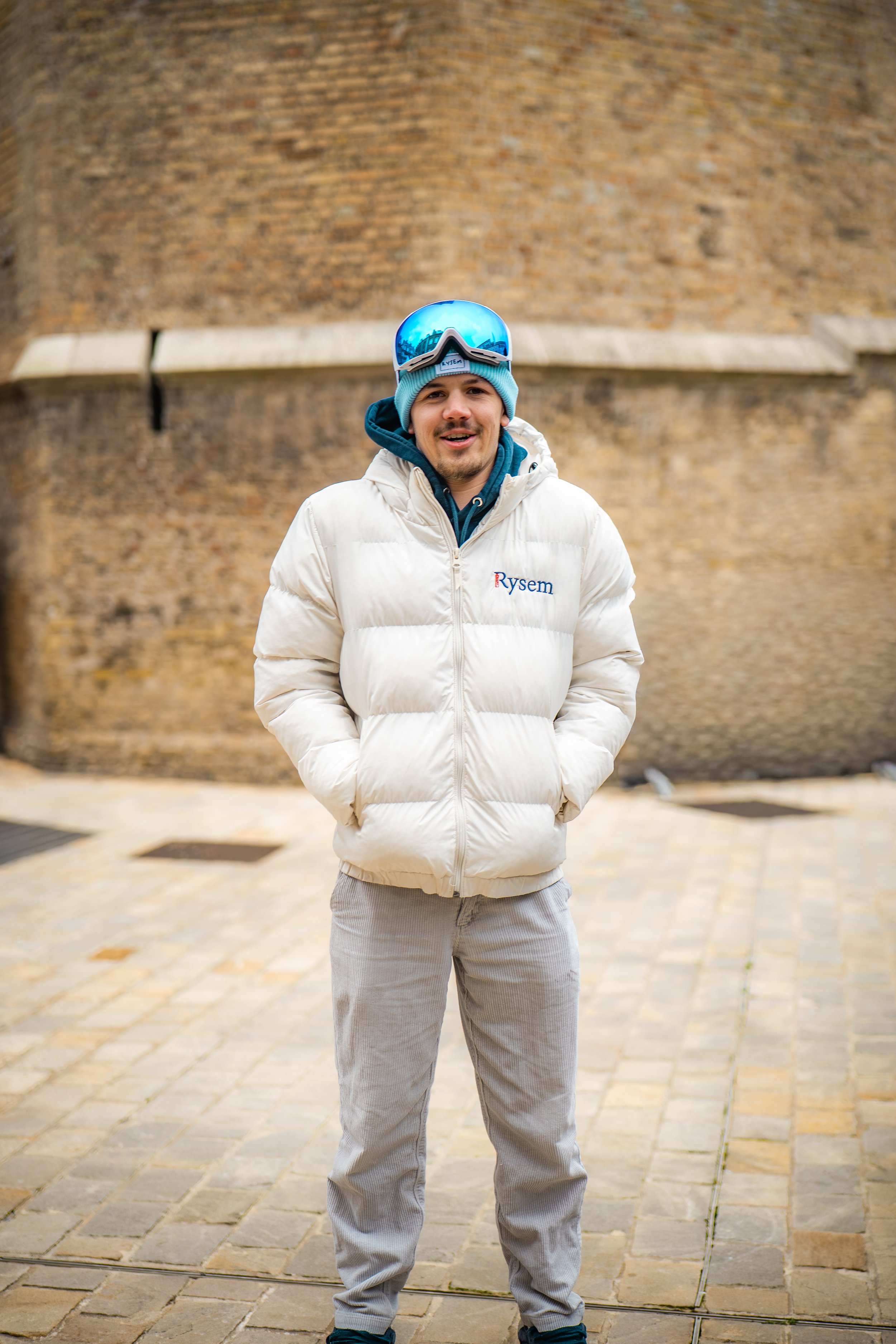 Un homme souriant portant une veste blanche, un bonnet bleu, des lunettes de ski bleues, et des pantalons gris, se tenant devant un fond en brique.