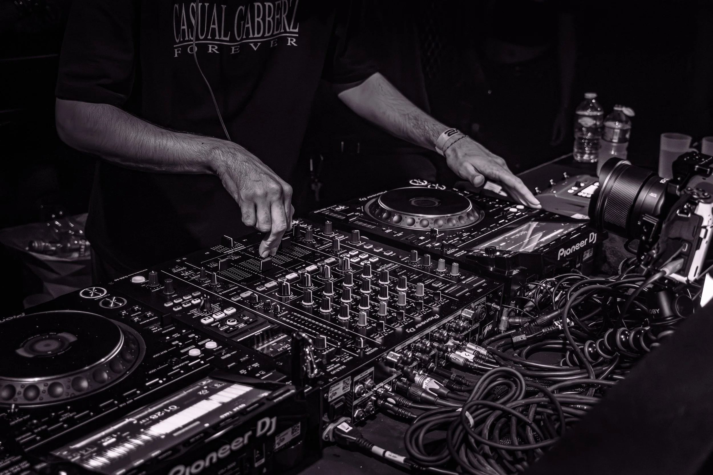 Une personne mixant de la musique avec un DJ set, en noir et blanc, à une table avec des équipements de DJ, sans visage visible, dans un environnement sombre.