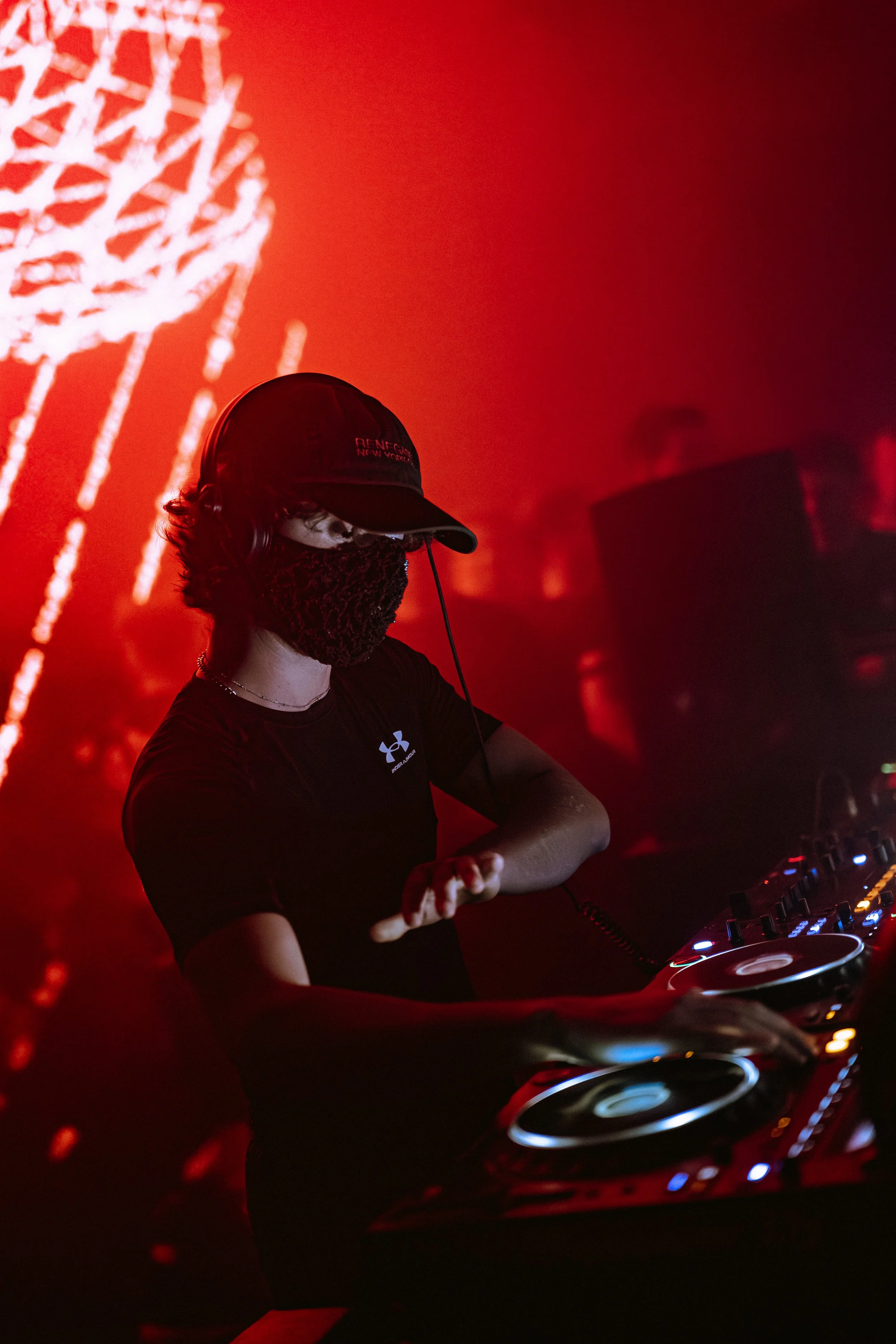 Un DJ portant un masque et une casquette noire, jouant de la musique dans une ambiance sombre et rouge lors d'une soirée.