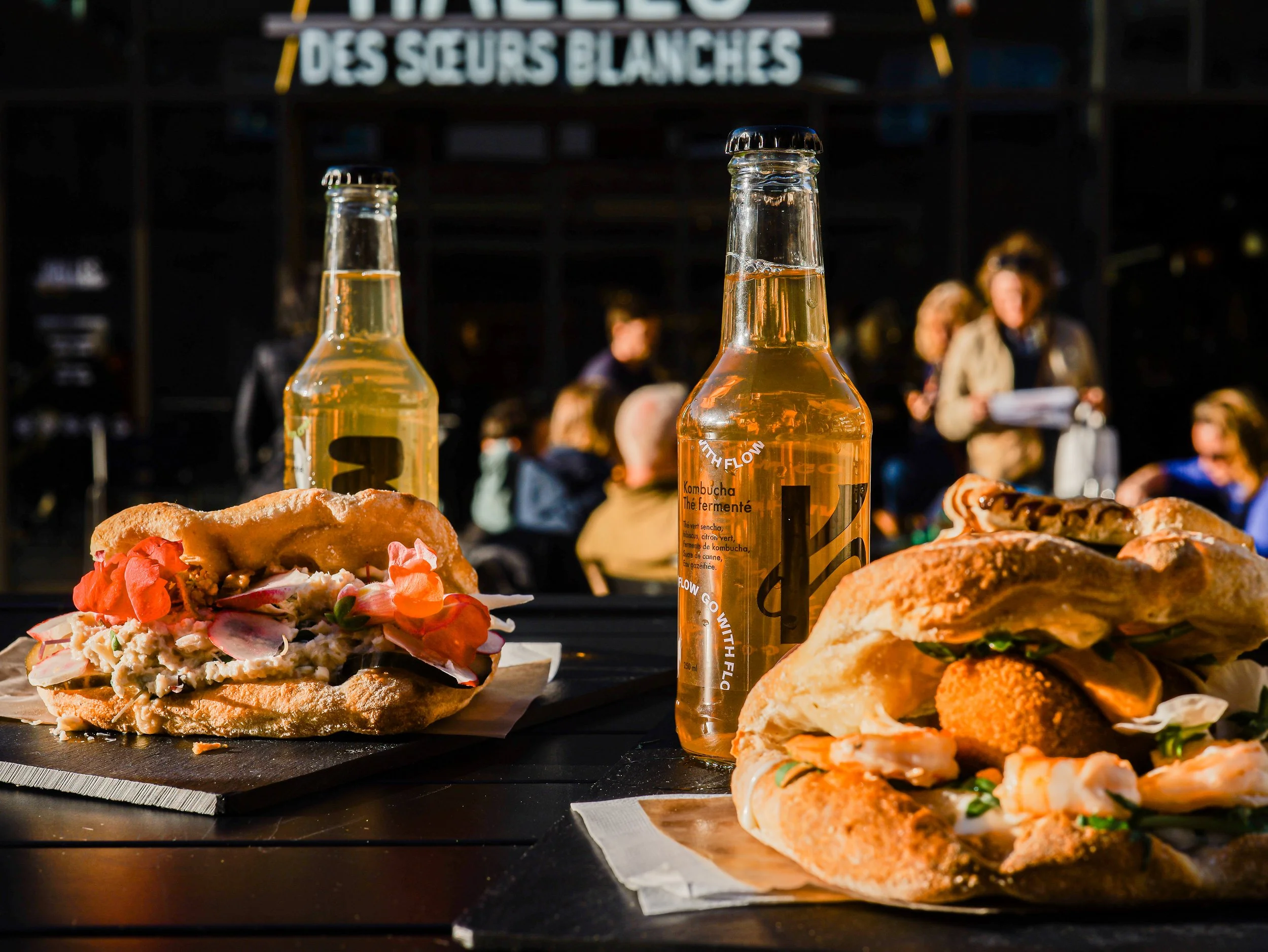 Bouteilles de boisson gazeuse sur une table, avec sandwichs et plats de nourriture, arrière-plan flou avec des personnes à un événement extérieur
