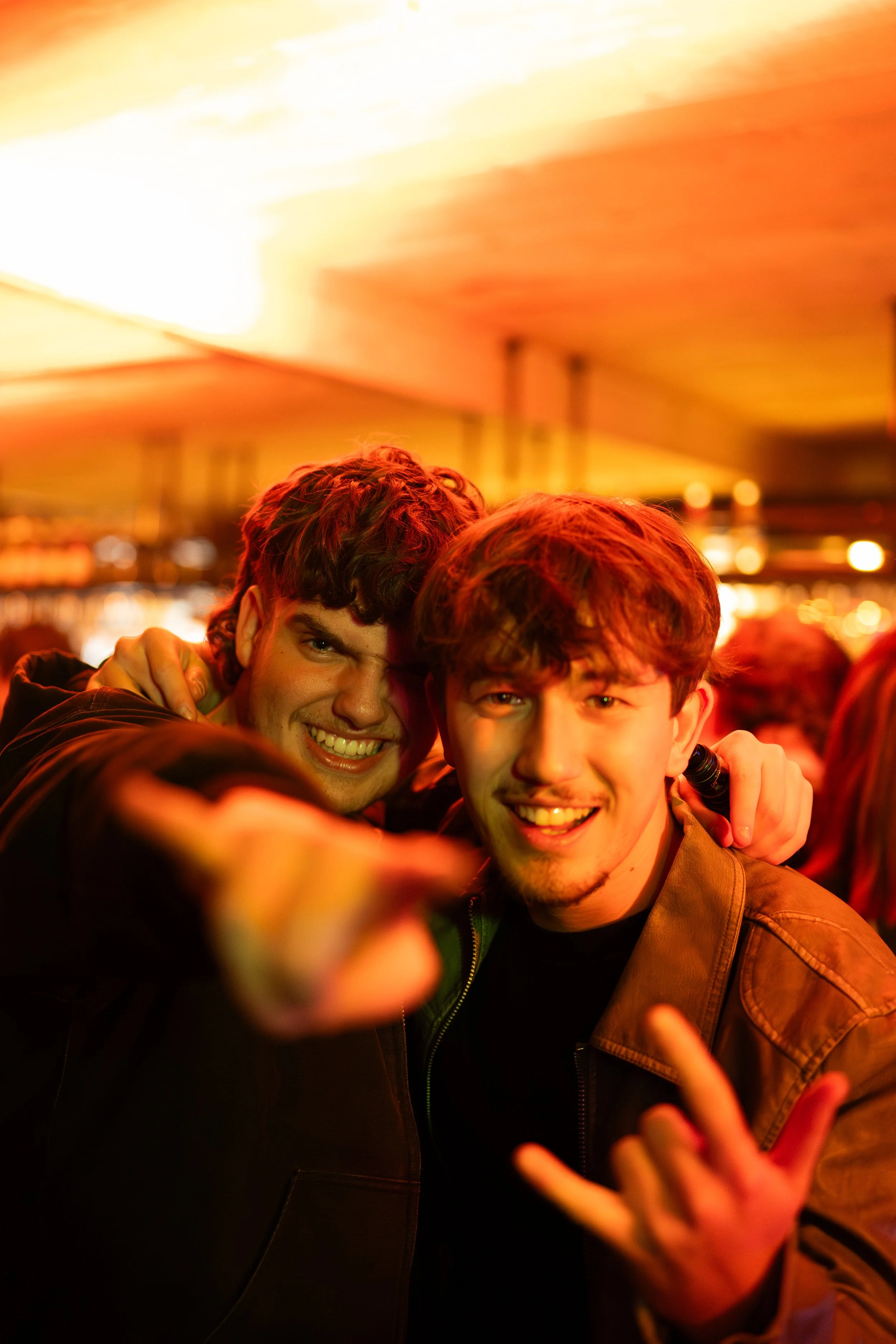 Deux jeunes hommes souriants posent pour une photo dans un environnement chaud et lumineux, avec une ambiance festive ou de fête.