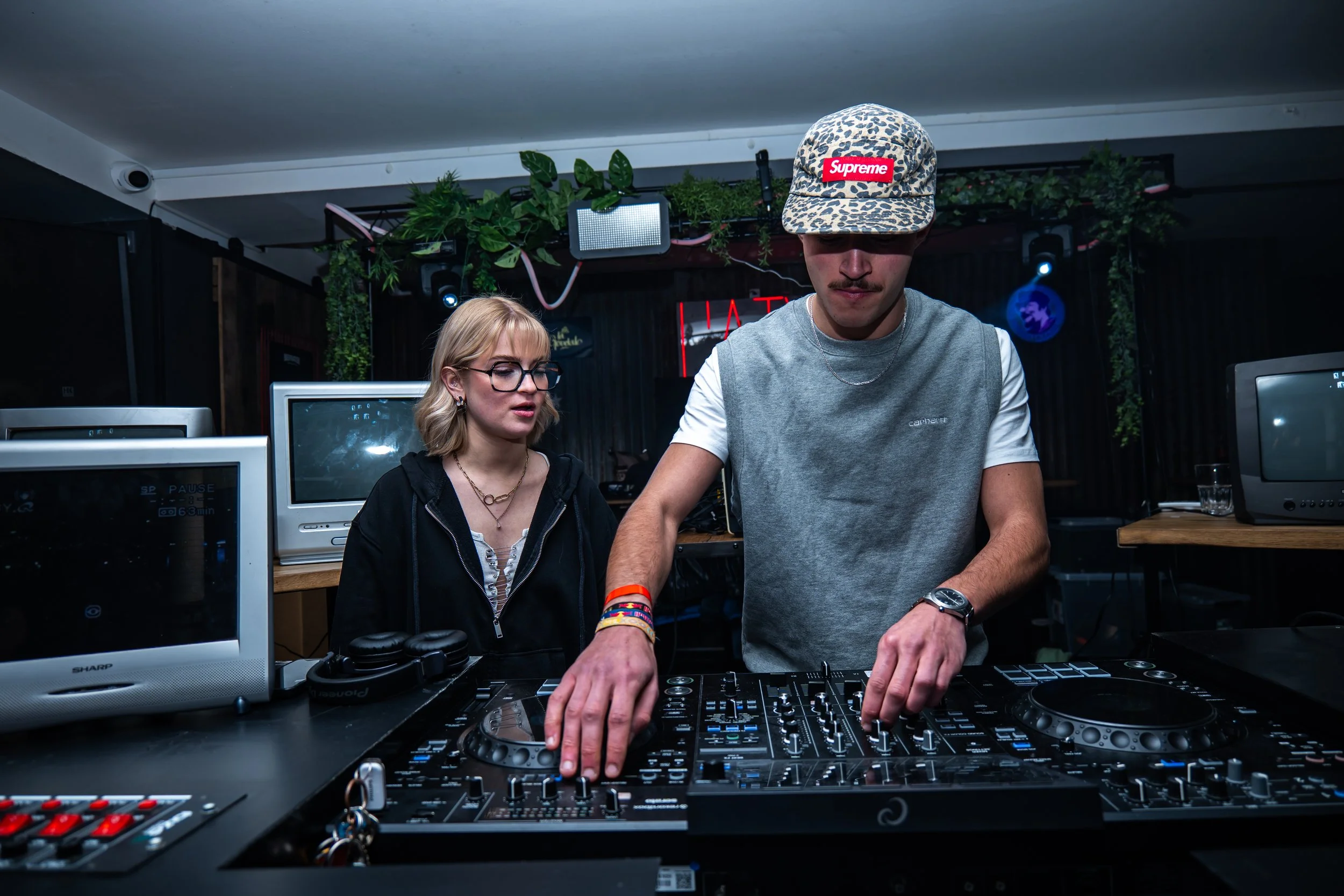 Un jeune homme portant une casquette à motif léopard avec un logo Supreme joue de la musique en tant que DJ, accompagné d'une femme portant des lunettes et un hoodie noir dans une ambiance sombre et urbaine avec des écrans et de la végétation en déco