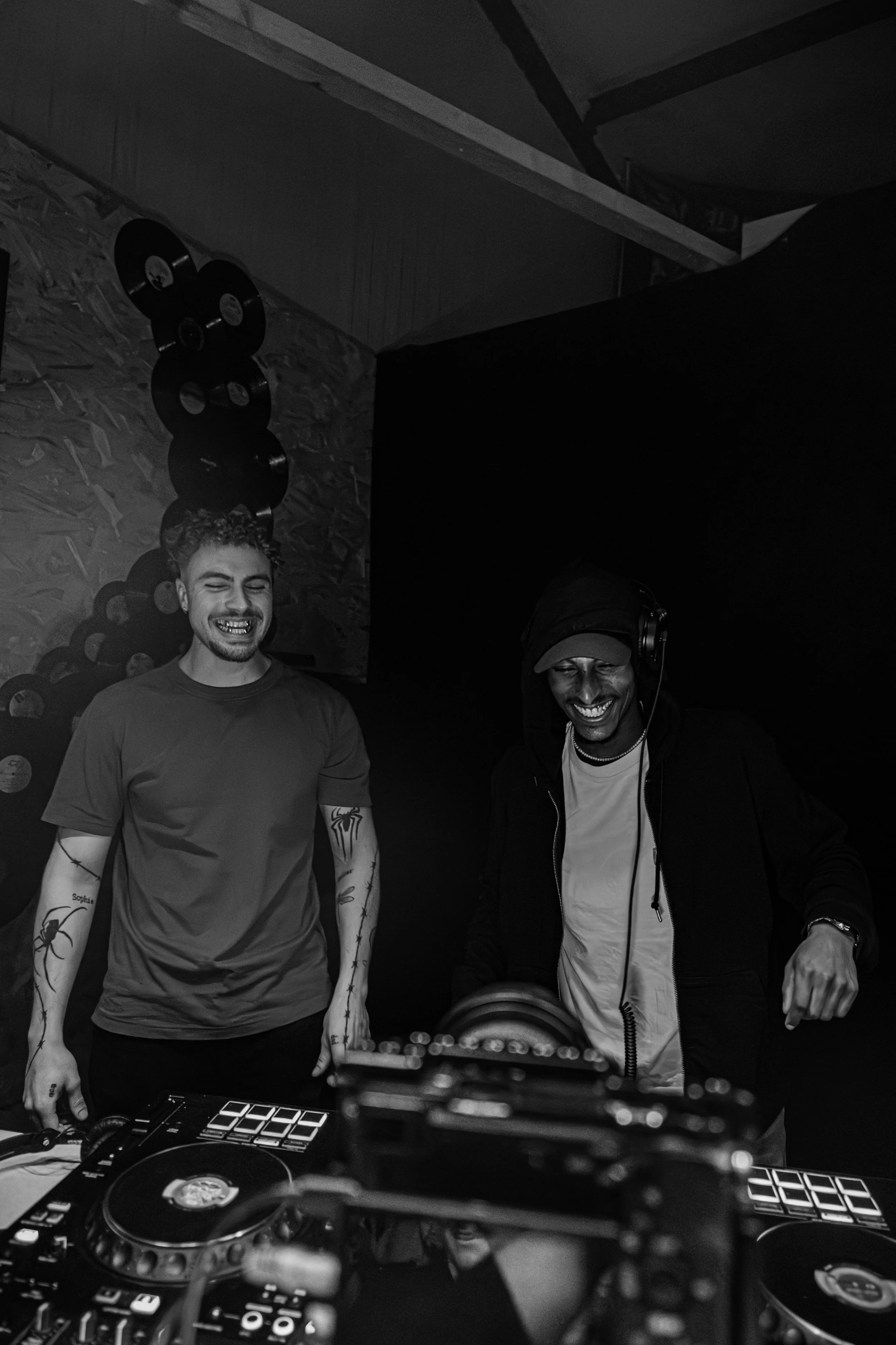 Deux hommes souriants derrière une table de DJ avec du matériel de musique, dans une pièce sombre