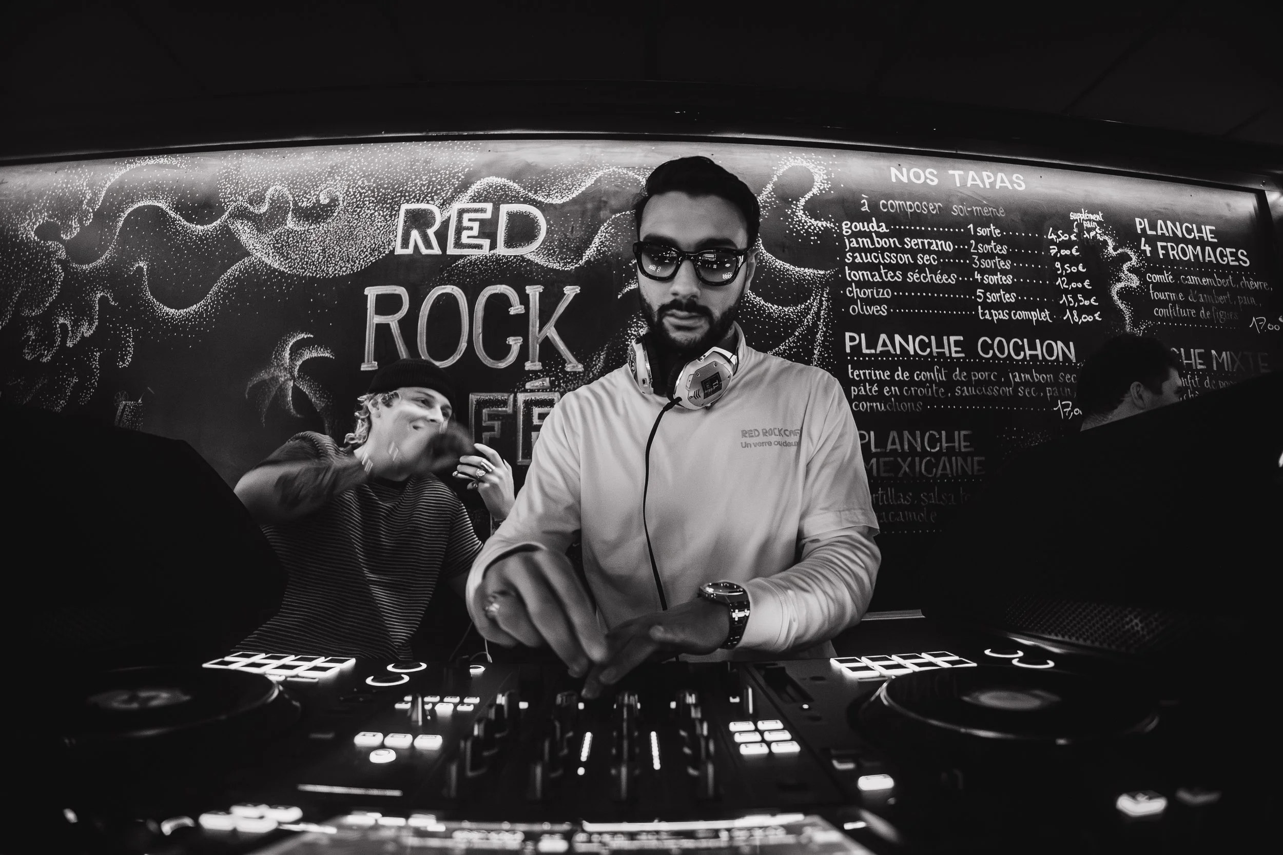 Un DJ avec lunettes de soleil derrière un platine, devant un tableau noir avec inscription "RED ROCK" et un menu en français.