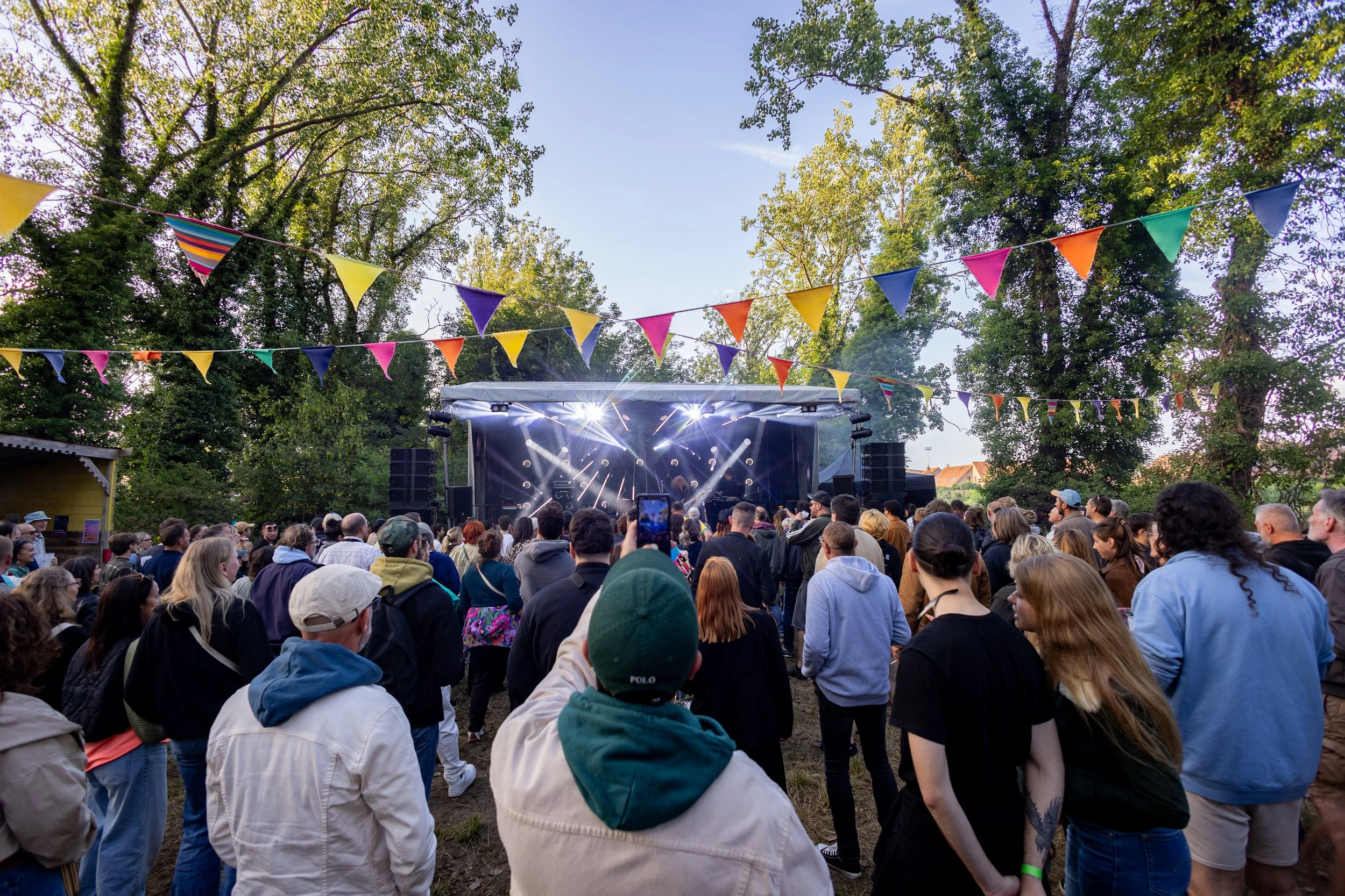 Une foule de personnes assises et debout dans un parc, regardant un spectacle sur scène. La scène est éclairée avec des lumières et décorée avec des guirlandes de fanions colorés suspendues entre les arbres. Le ciel est clair et le spectacle semble s
