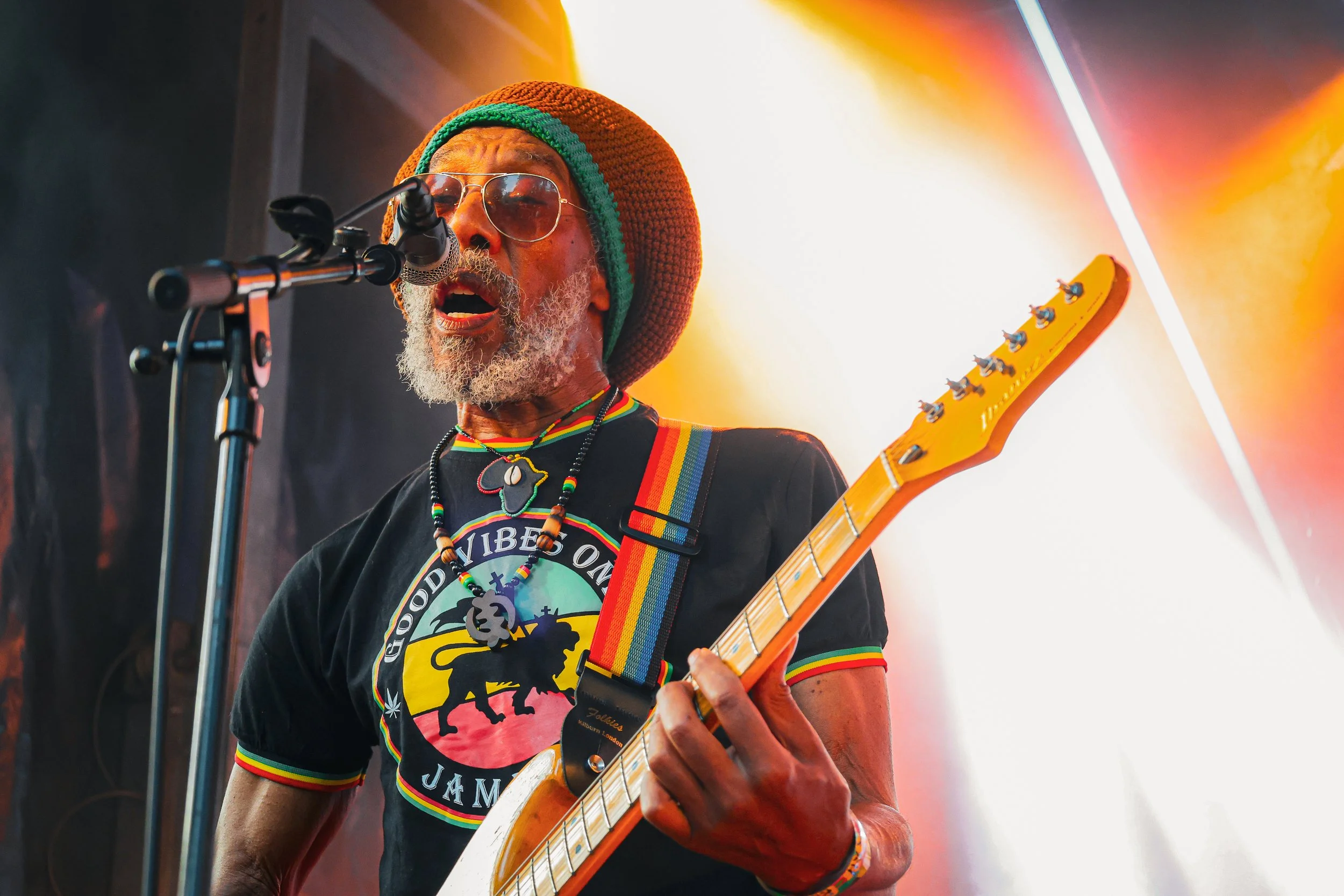 Un musicien afro-américain portant un chapeau en tricot orange, des lunettes de soleil, un t-shirt avec un logo coloré, et jouant de la guitare électrique lors d'un concert. Il chante dans un microphone avec un fond lumineux orange et blanc.