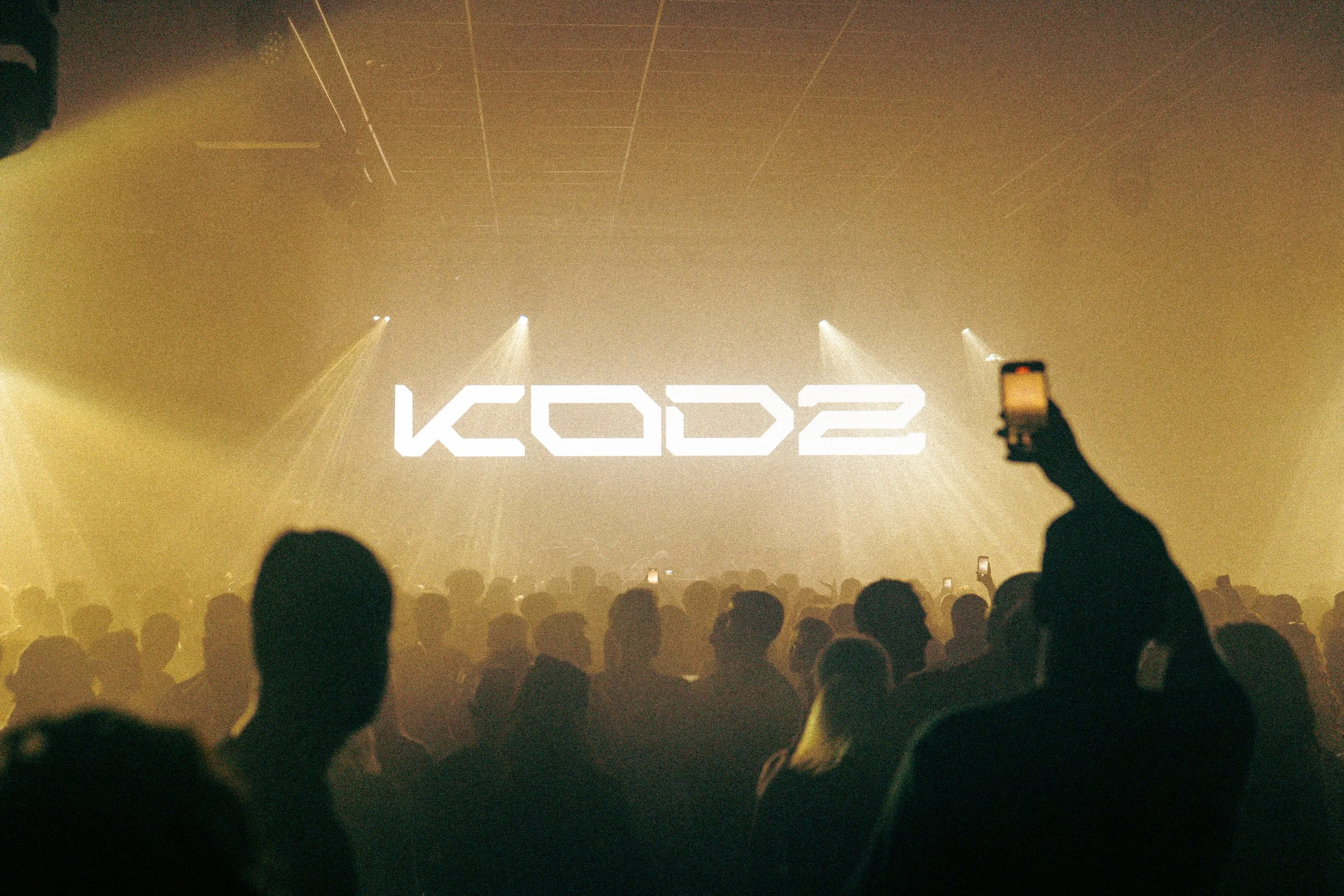 Concert avec un grand écran affichant le texte 'KOD' en lettres blanches. La scène est éclairée de lumières jaunes, et une foule regarde le spectacle, certains prenant des photos avec leur téléphone.