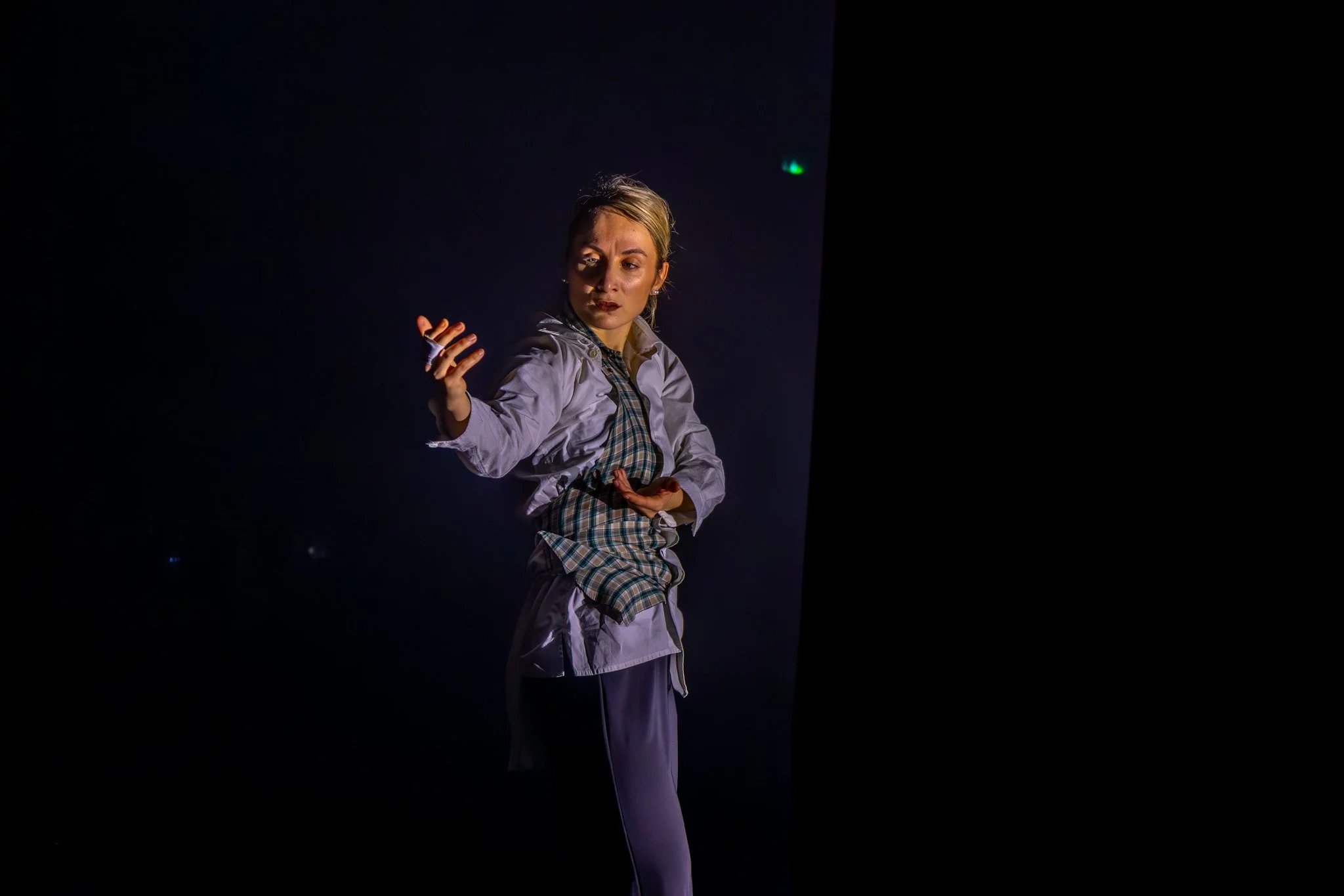 Une femme danse dans une scène sombre, portant une tenue de danse avec un haut gris, un pantalon gris et une veste à carreaux. Elle a les cheveux attachés en arrière et exprime une posture expressive.