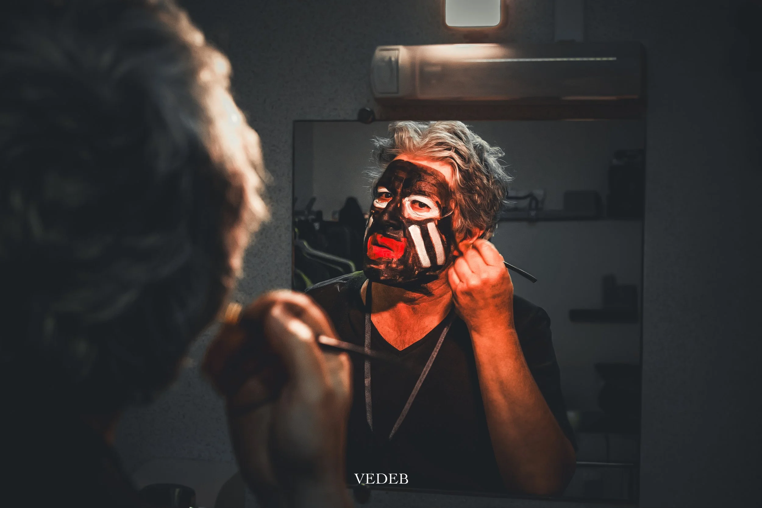 Une personne se maquille devant un miroir dans une pièce sombre. Elle a les yeux maquillés et une peinture sur le visage, avec une expression concentrée.