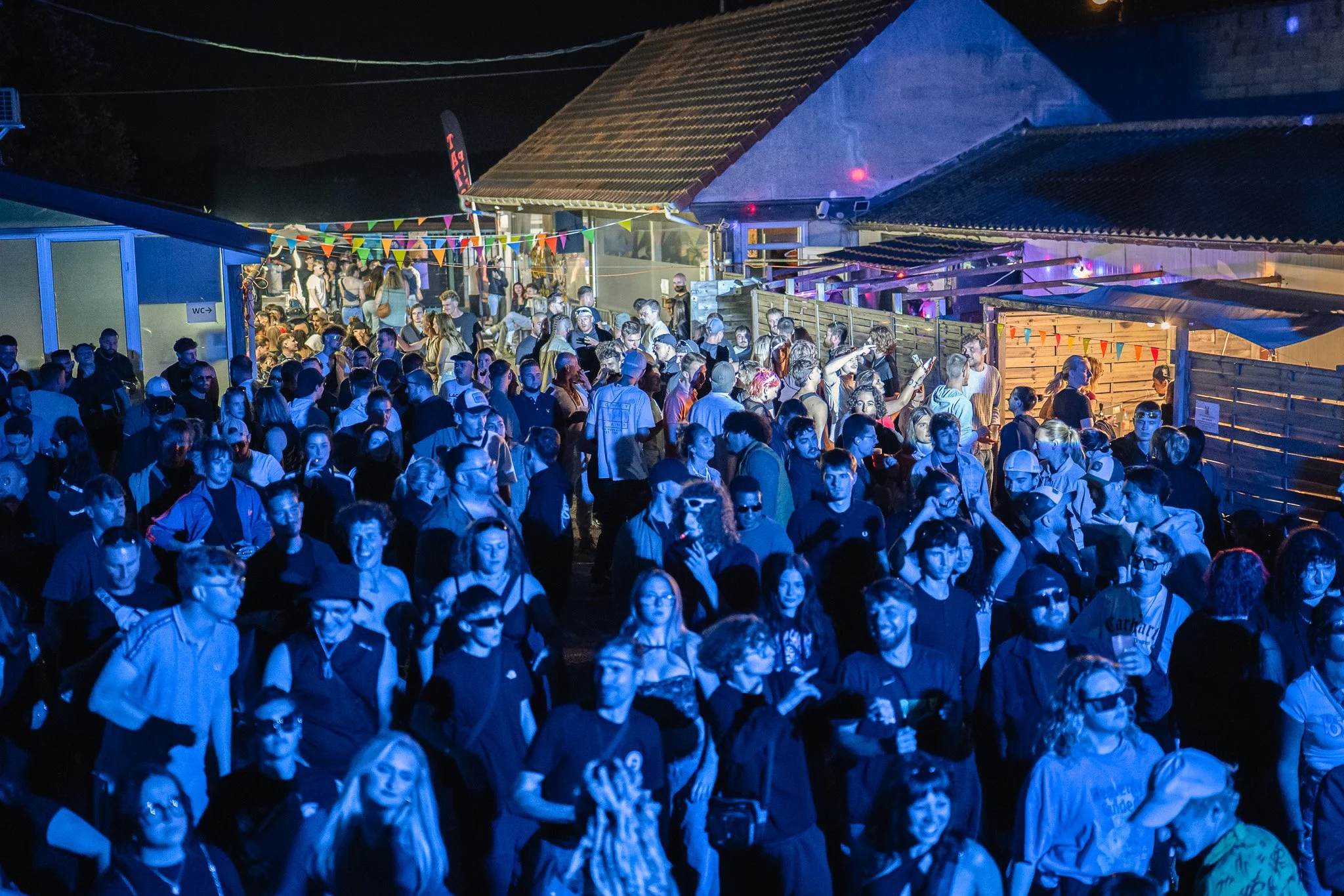 Une foule de personnes lors d'un événement nocturne en extérieur, avec des décorations colorées et un bâtiment en arrière-plan.