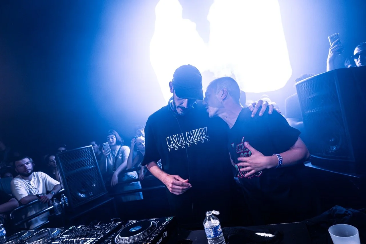 Deux personnes discutent étroitement derrière un DJ sur une scène dans un club, avec plusieurs spectateurs en arrière-plan et un éclairage bleu vif.