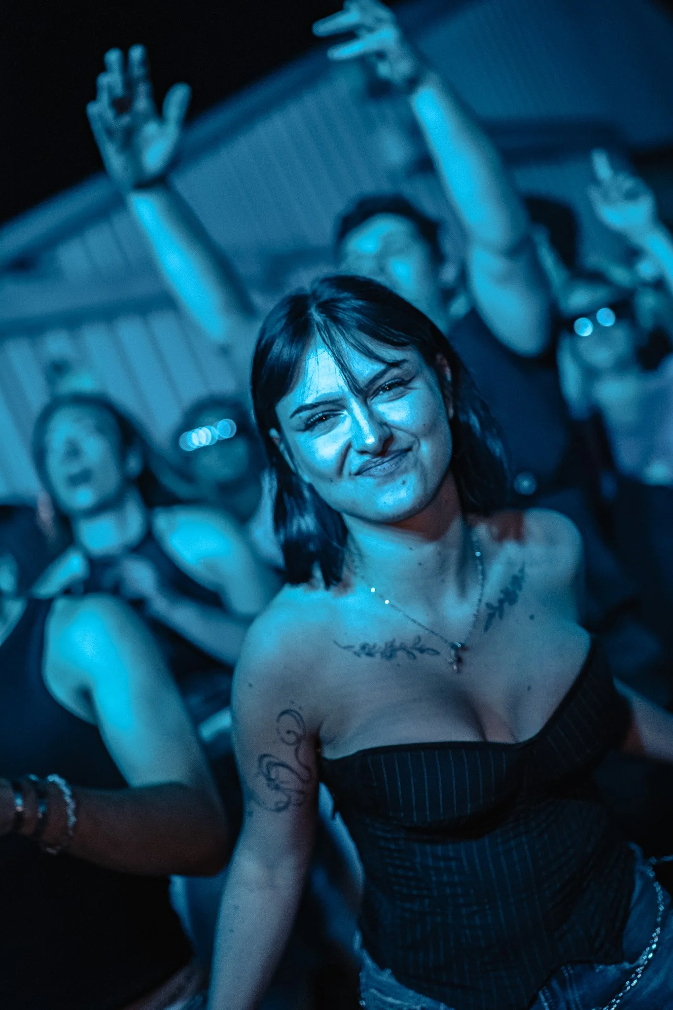 Personne souriante avec des tatouages, portant une robe à rayures, entourée d'autres personnes lors d'une fête ou événement, sous musique ou lumière bleue, ambiance joyeuse.