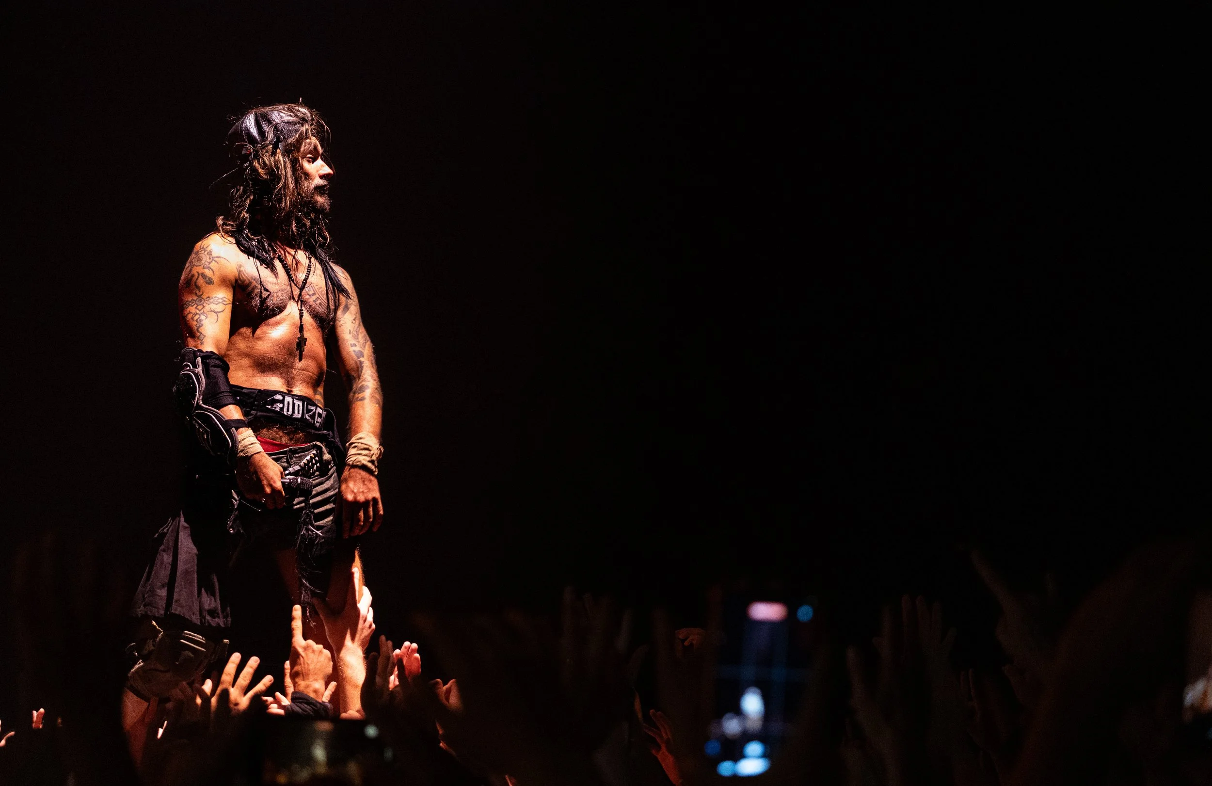 Un homme avec des tatouages et des dreadlocks, probablement un musicien, se tient sur scène, salué par un public dont les mains sont levées.