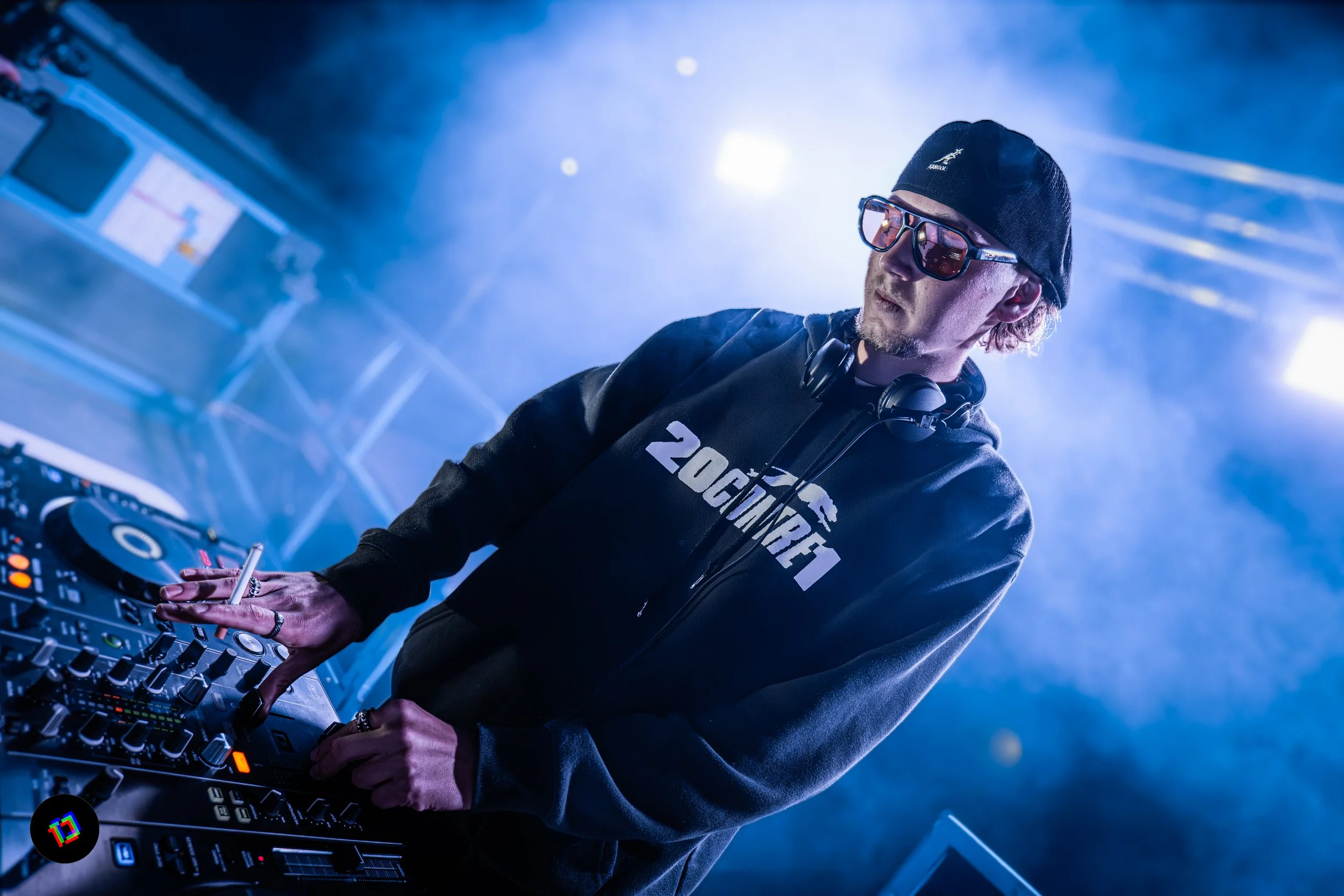 DJ mixant des chansons dans une scène lumineuse de concert ou de club, portant des lunettes de soleil et un bonnet noir.