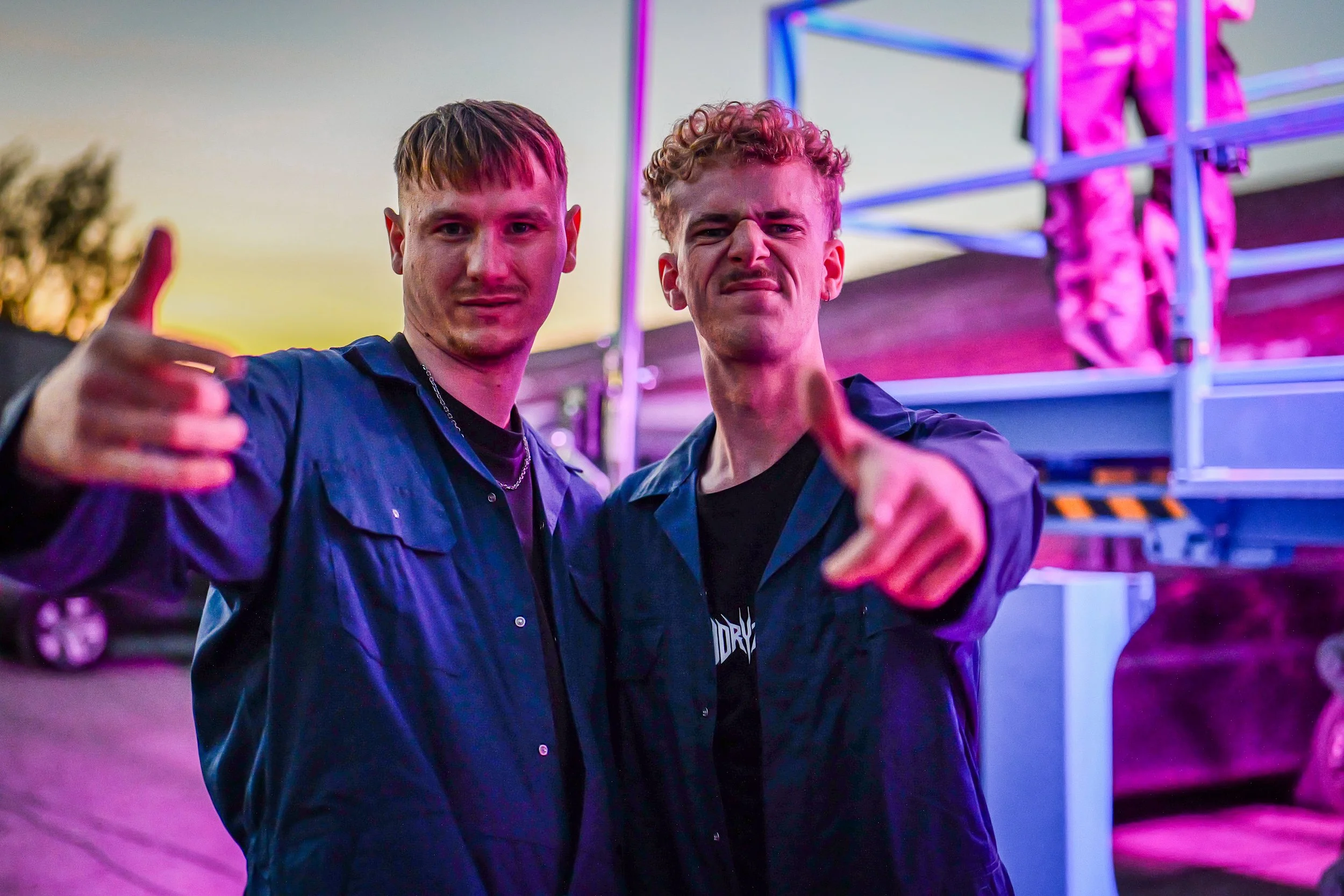 Deux jeunes hommes posent pour une photo lors d'un événement nocturne, avec un éclairage néon coloré en arrière-plan.