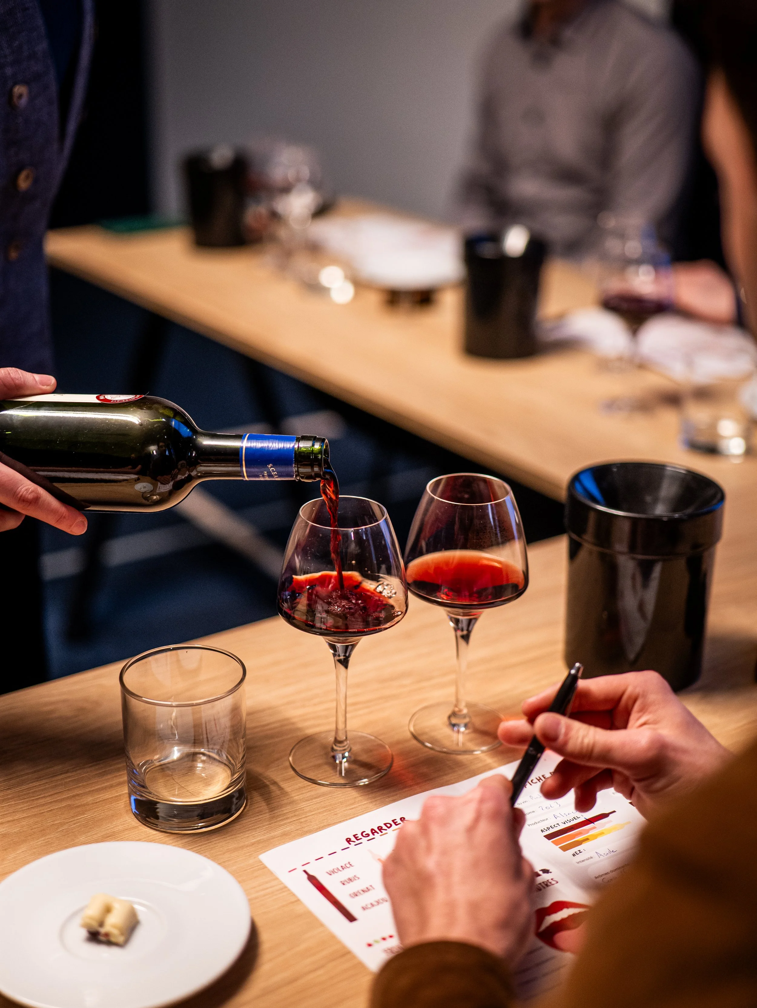 Personne versant du vin rouge dans un verre lors d'une dégustation, avec d'autres verres, une fiche d'évaluation, et un plateau de fromage sur la table.