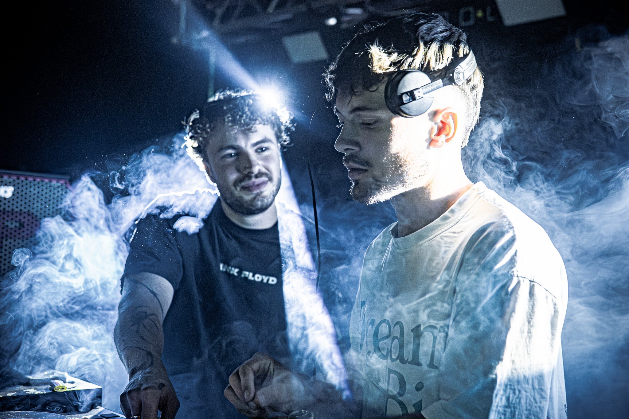 Deux jeunes hommes en studio, portant des casques audio, entourés de fumée et de lumières colorées, semblent en train de mixer de la musique.