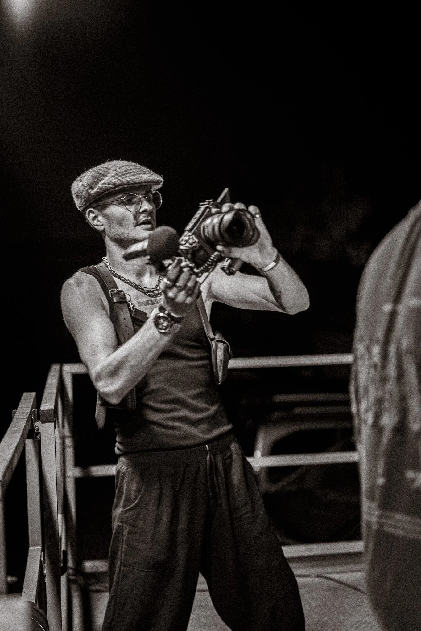 Un homme avec une casquette en tweed, lunettes, un maillot sans manches, une chaîne métallique, et des accessoires, tient une caméra professionnelle dans ses mains, en train de filmer ou prendre en photo.