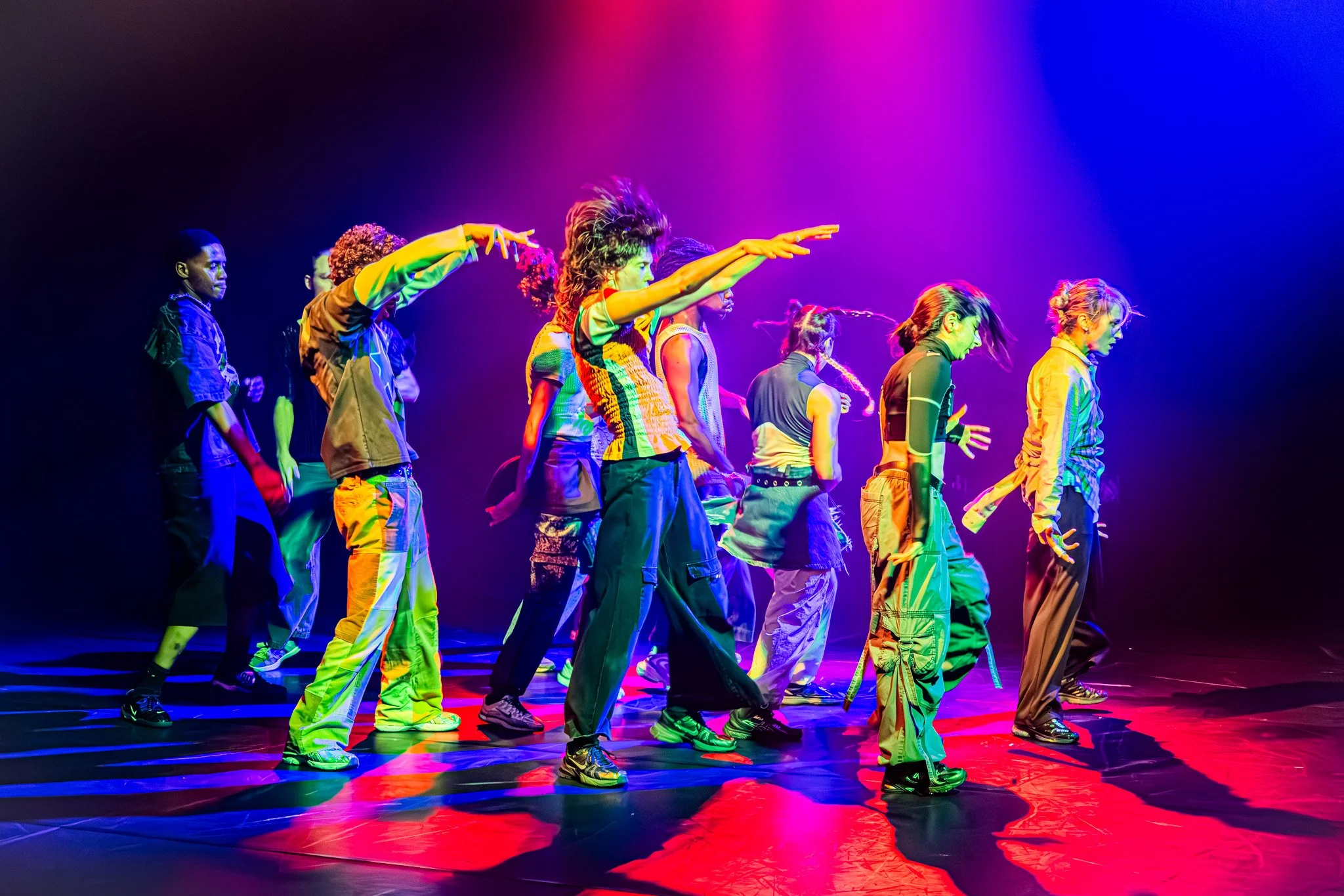 Groupe de danseurs colorés en mouvement sous des lumières violettes et rouges dans une scène de spectacle.