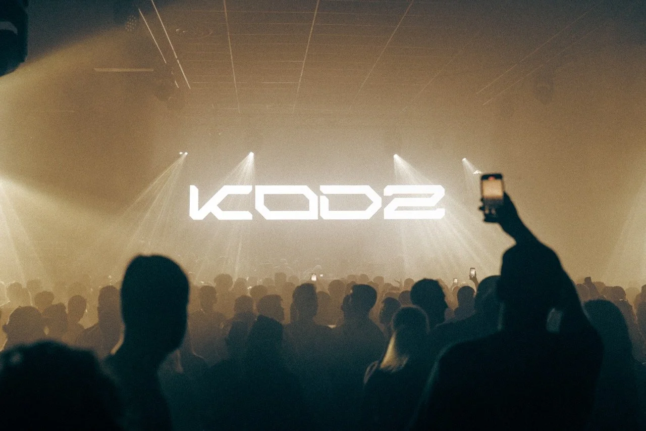 Une scène de concert avec de nombreuses personnes dans une salle sombre, éclairée par des lumières de scène. Le mot "KOD2" est affiché en grand à l'arrière-plan.