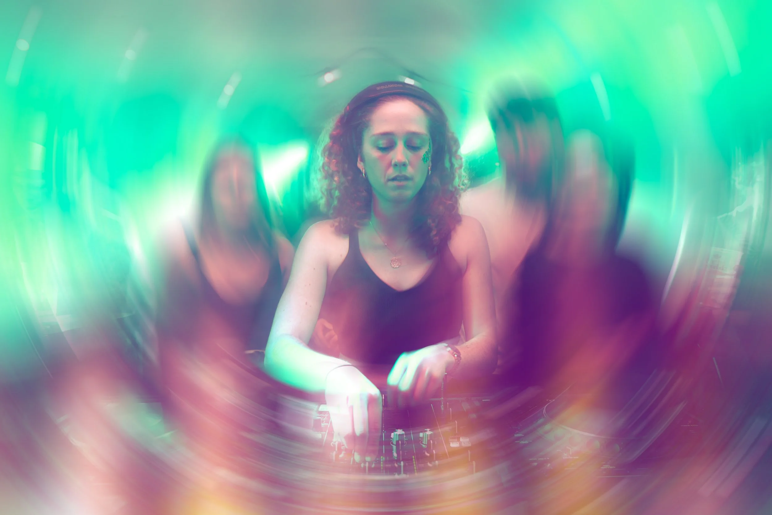 Une femme DJ avec des cheveux bouclés et un maquillage artistique, utilisant des écouteurs, dans une ambiance colorée et lumineuse lors d'une performance musicale.
