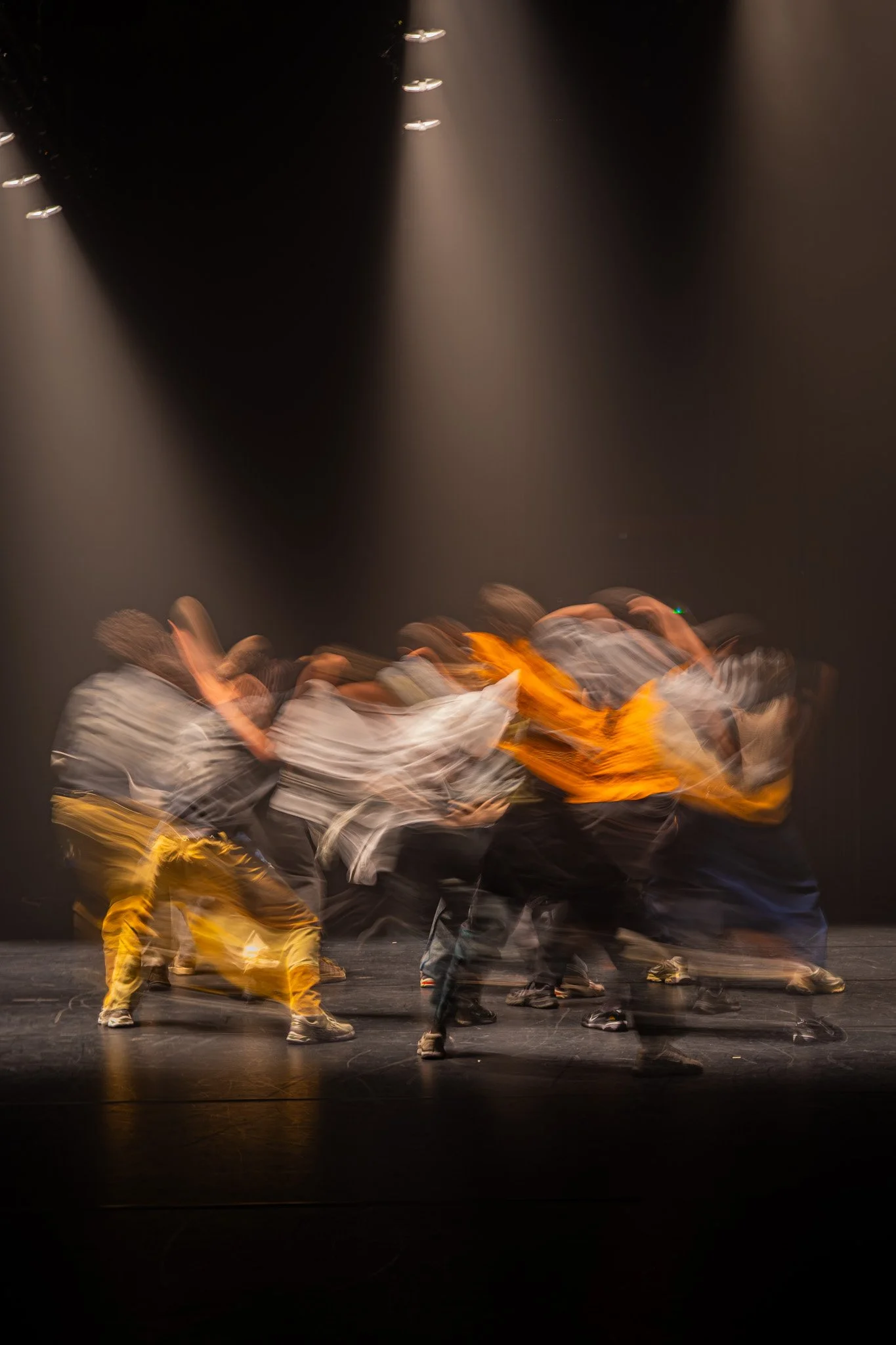 Groupe de danseurs en mouvement sur scène, éclairage dramatique, arrière-plan noir, vêtements décontractés avec couleurs vives, scène de performance dynamique.