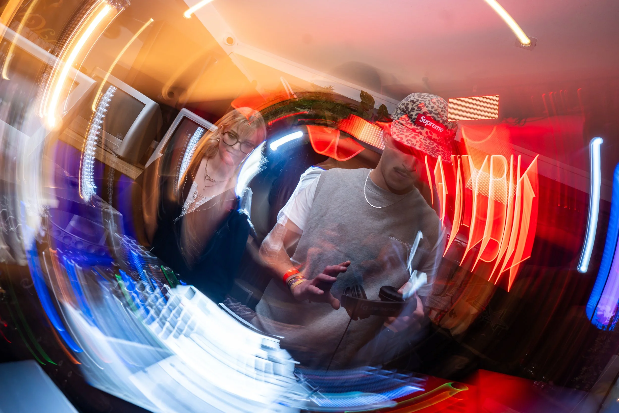 Deux personnes, un homme et une femme, DJ, en train de mixer dans un club ou un événement, avec des lumières colorées et des effets de mouvement.
